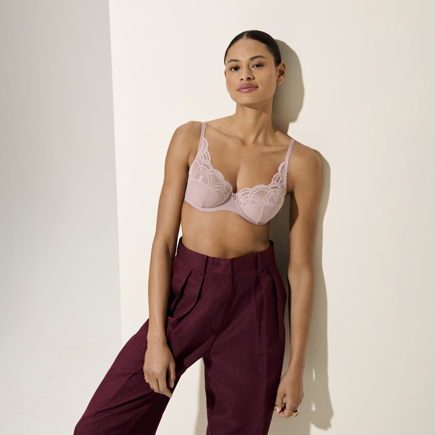 Woman styled in mauve lace underwire bra and burgundy trousers, posing against a neutral background.