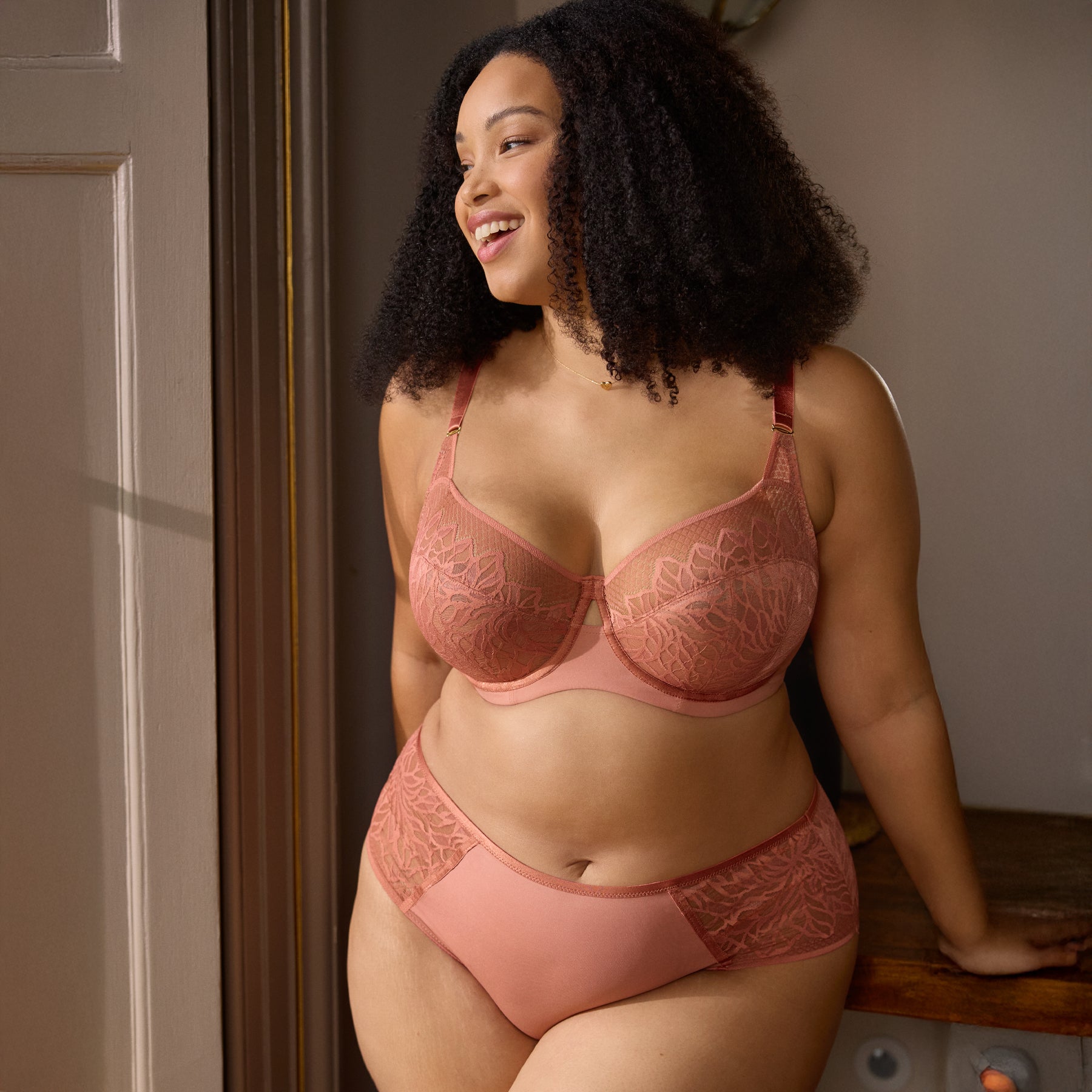 Woman wearing sienna rose lace bra and matching high-waist brief, smiling by a window.