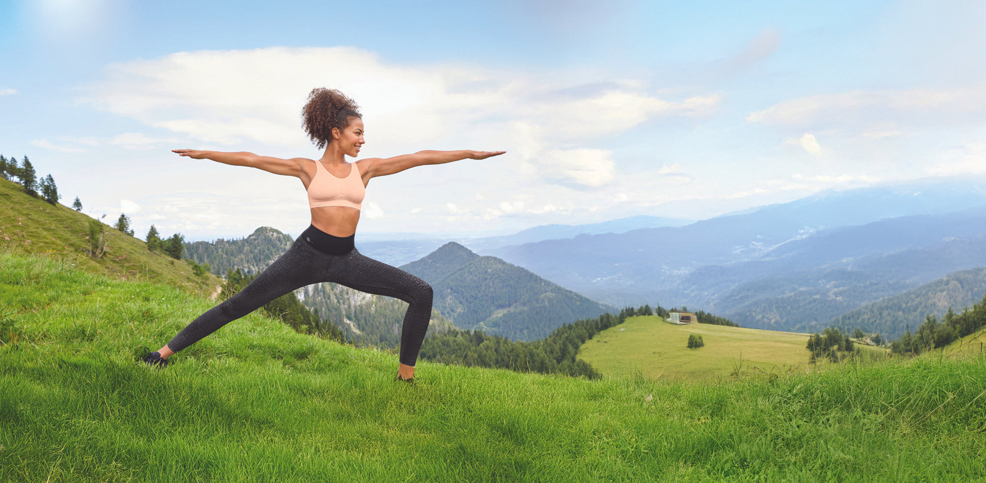 Woman practising yoga in the mountains wearing a blush pink Anita sports bra and high-rise leggings, surrounded by scenic alpine landscape
