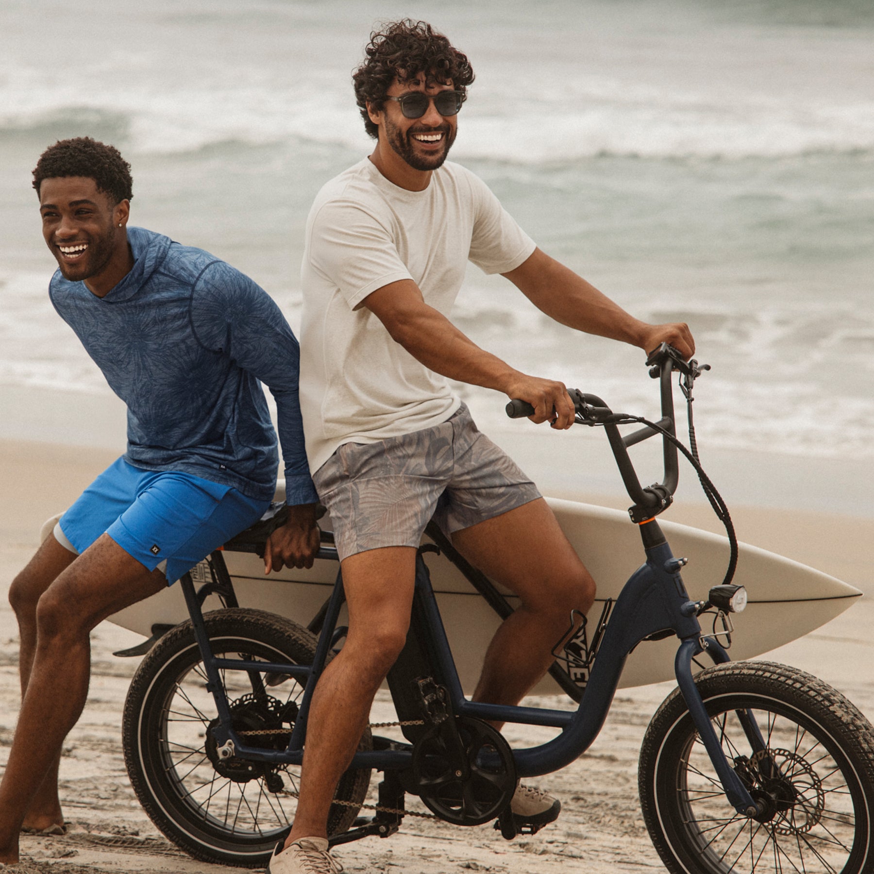 Man riding an electric bike on the beach with a friend, both wearing casual tops and swim shorts, smiling beside ocean waves and a surfboard