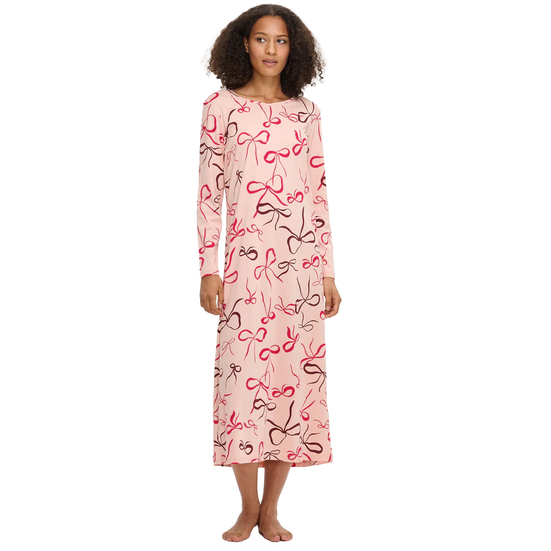 Woman wearing a long pink nightdress with red and brown bow print, long sleeves, and a relaxed fit, standing barefoot against a white background.