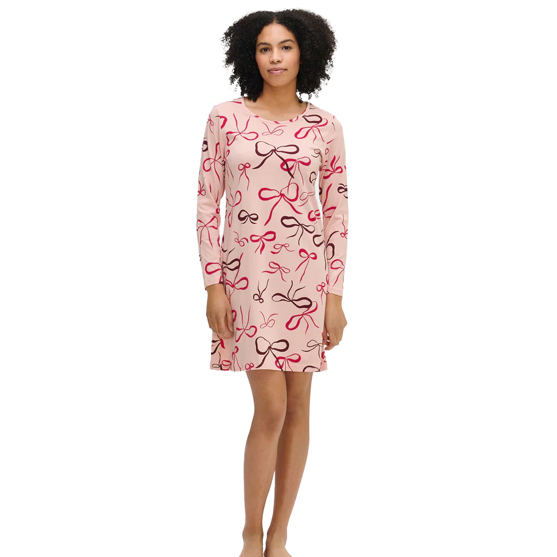 Woman wearing a long-sleeve pink nightdress with red and brown bow print, standing barefoot against a white background.