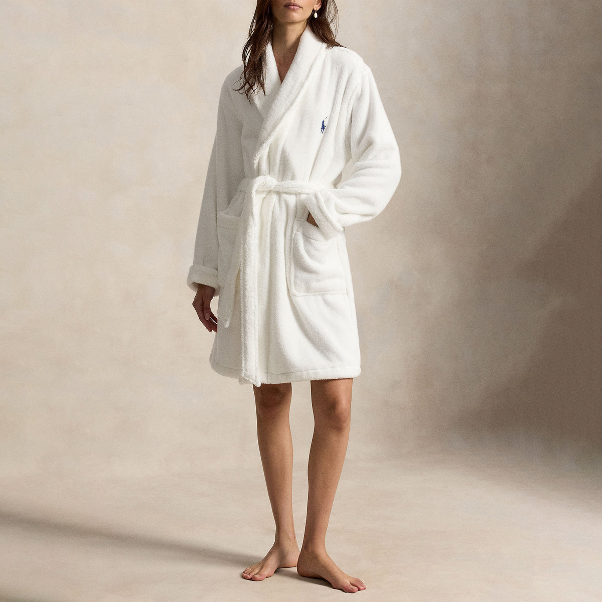 Model wearing a white Polo Ralph Lauren plush fleece robe featuring a shawl collar, adjustable tie belt, front patch pockets, and signature navy Polo logo.
