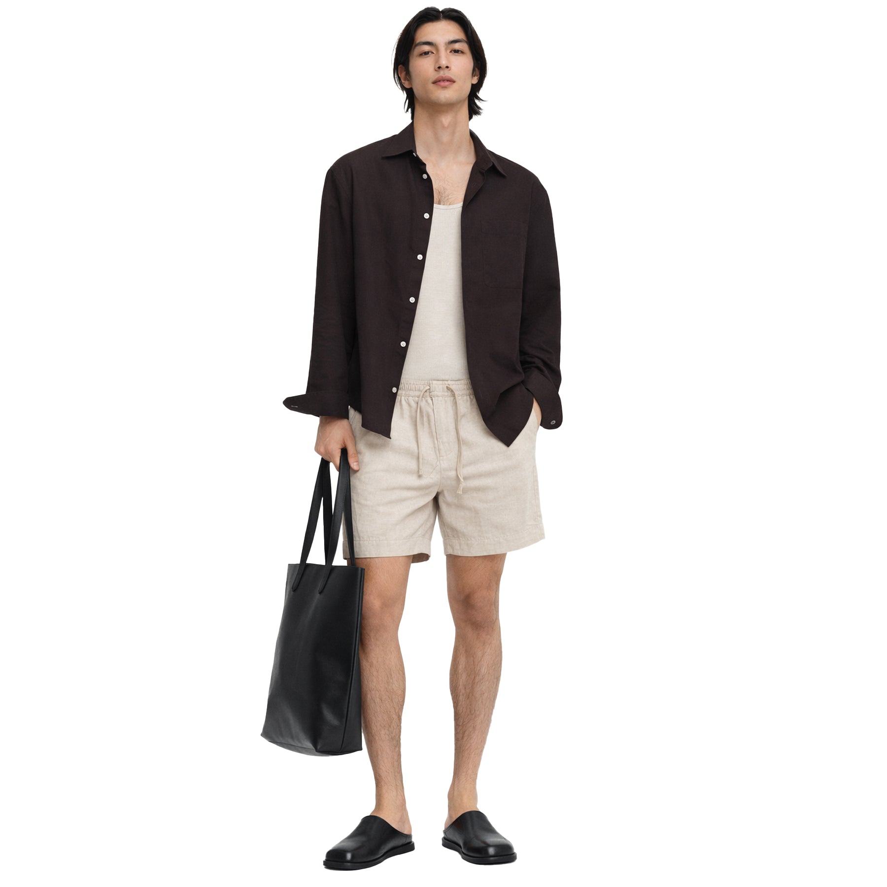 Male model wearing beige linen drawstring shorts styled with a neutral tank, dark shirt and black slip-on shoes.