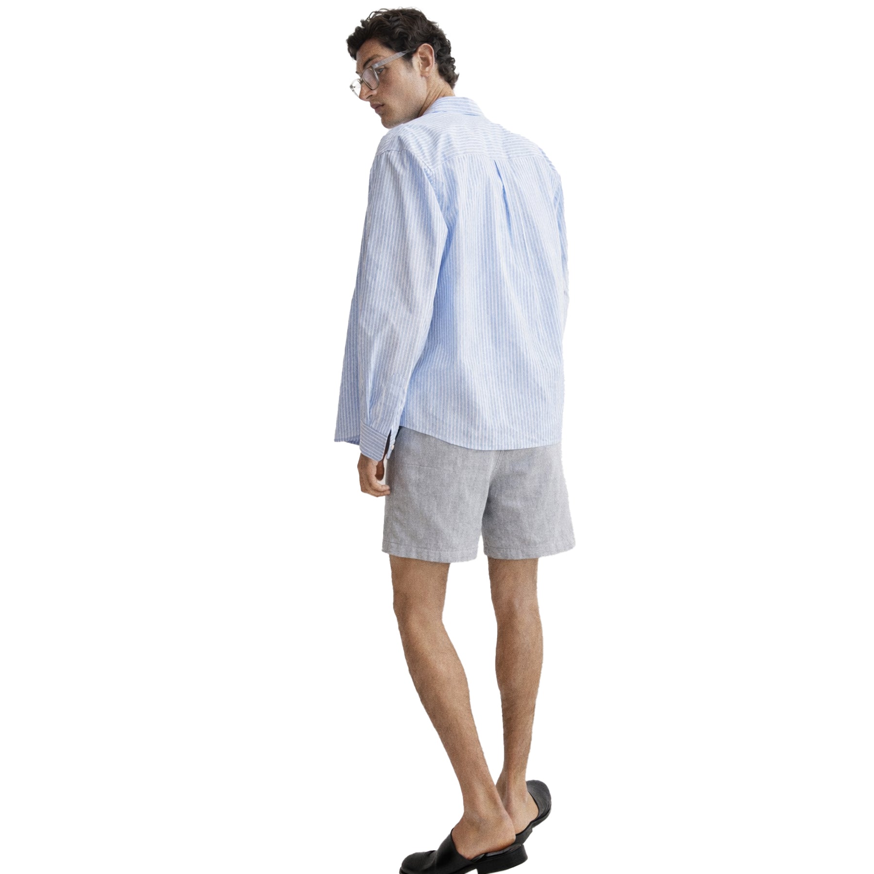 Male model wearing light grey linen shorts styled with a blue striped shirt and black slip-on shoes, back view.