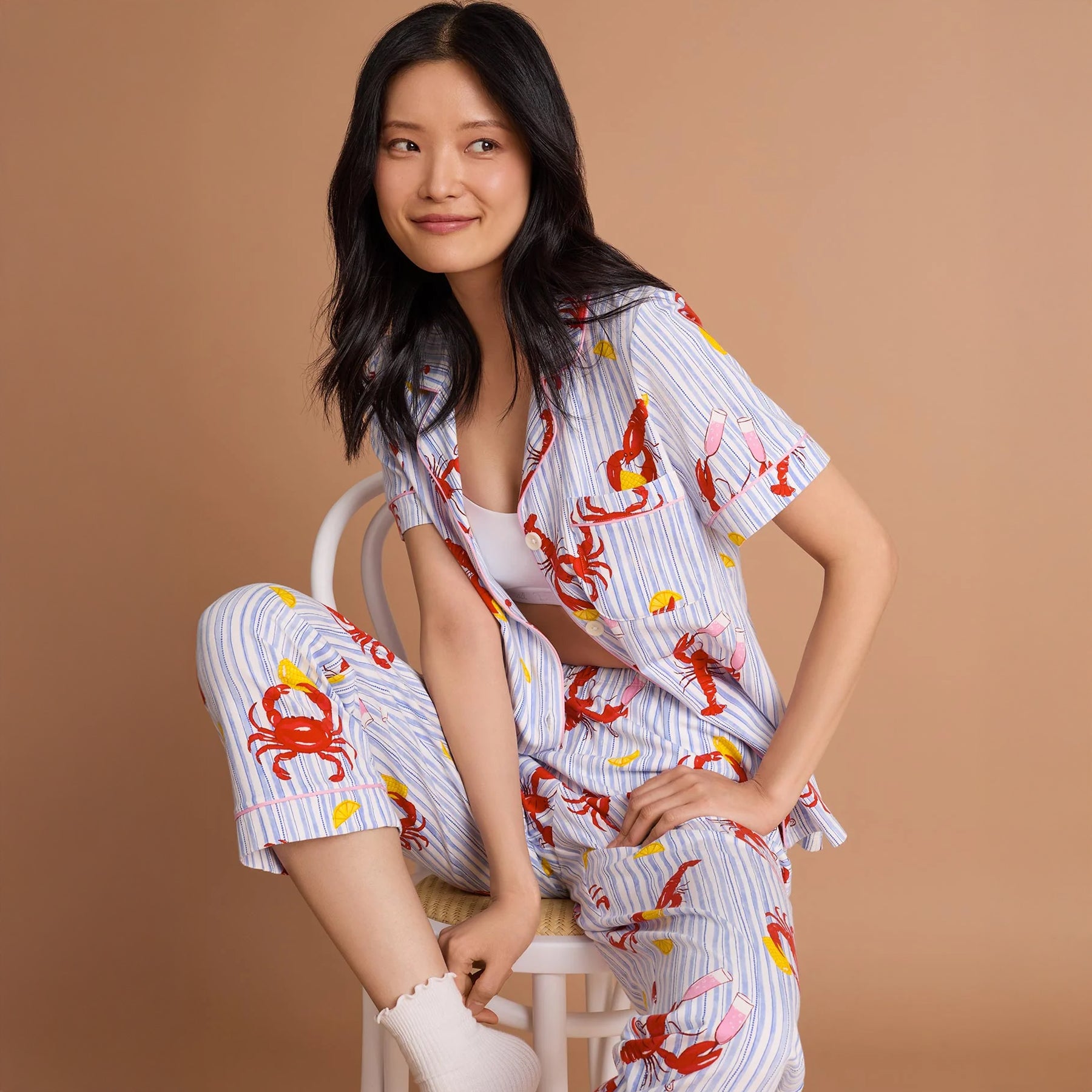Woman wearing a blue and white striped pyjama set with playful red lobster and crab print, featuring a short sleeve button-front top and cropped pants.