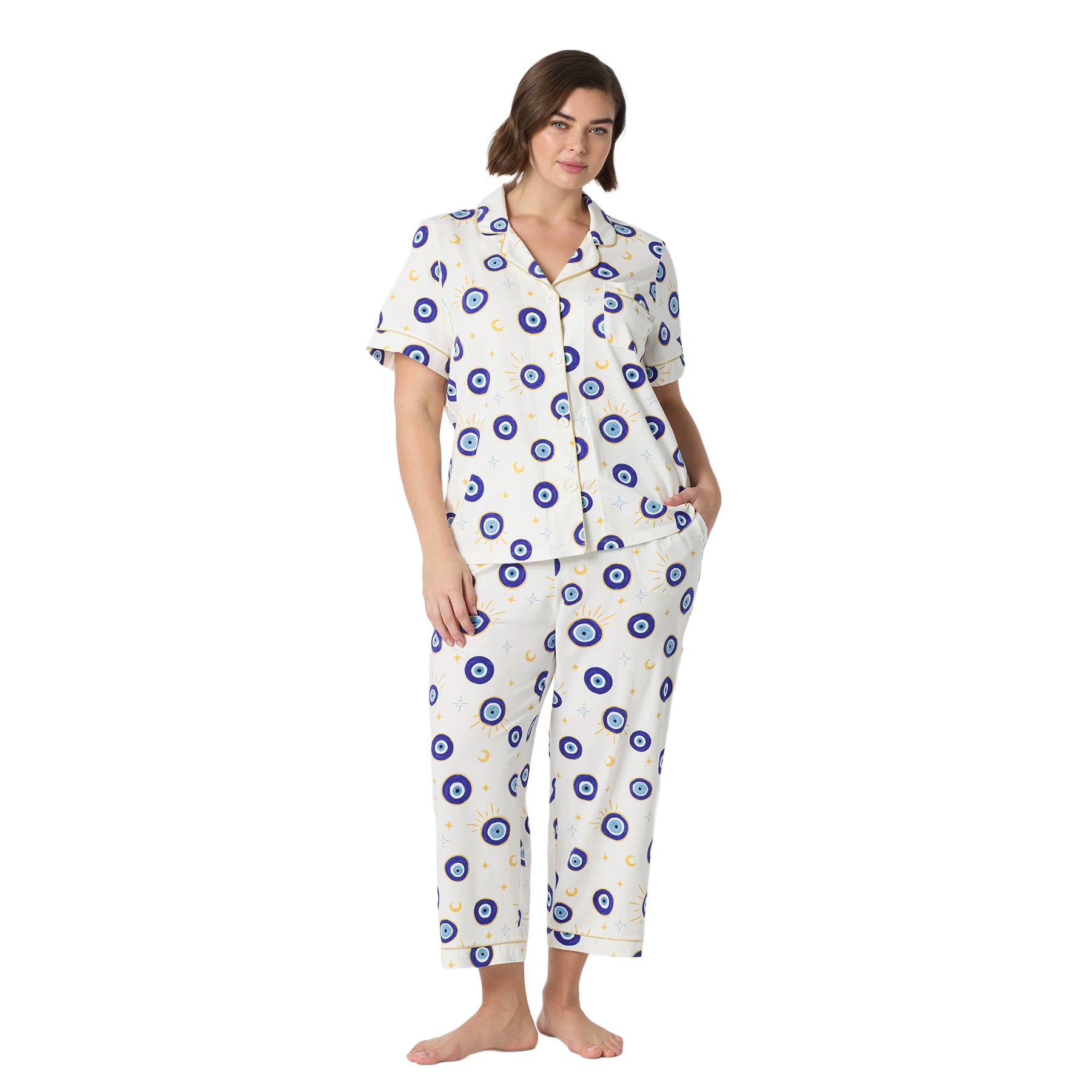 Plus-size woman wearing an ivory short-sleeve pajama set with cropped pants, blue evil eye celestial print and gold piping.