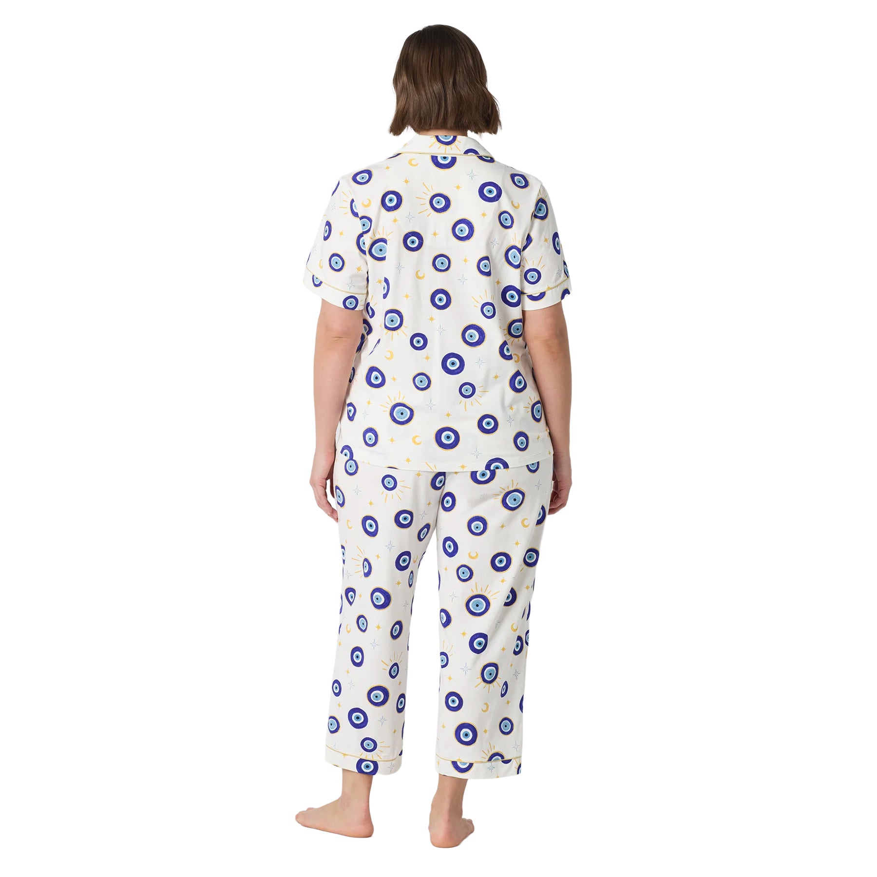 Back view of a plus-size woman wearing an ivory evil eye print pajama set with short sleeves, cropped pants and gold piping.