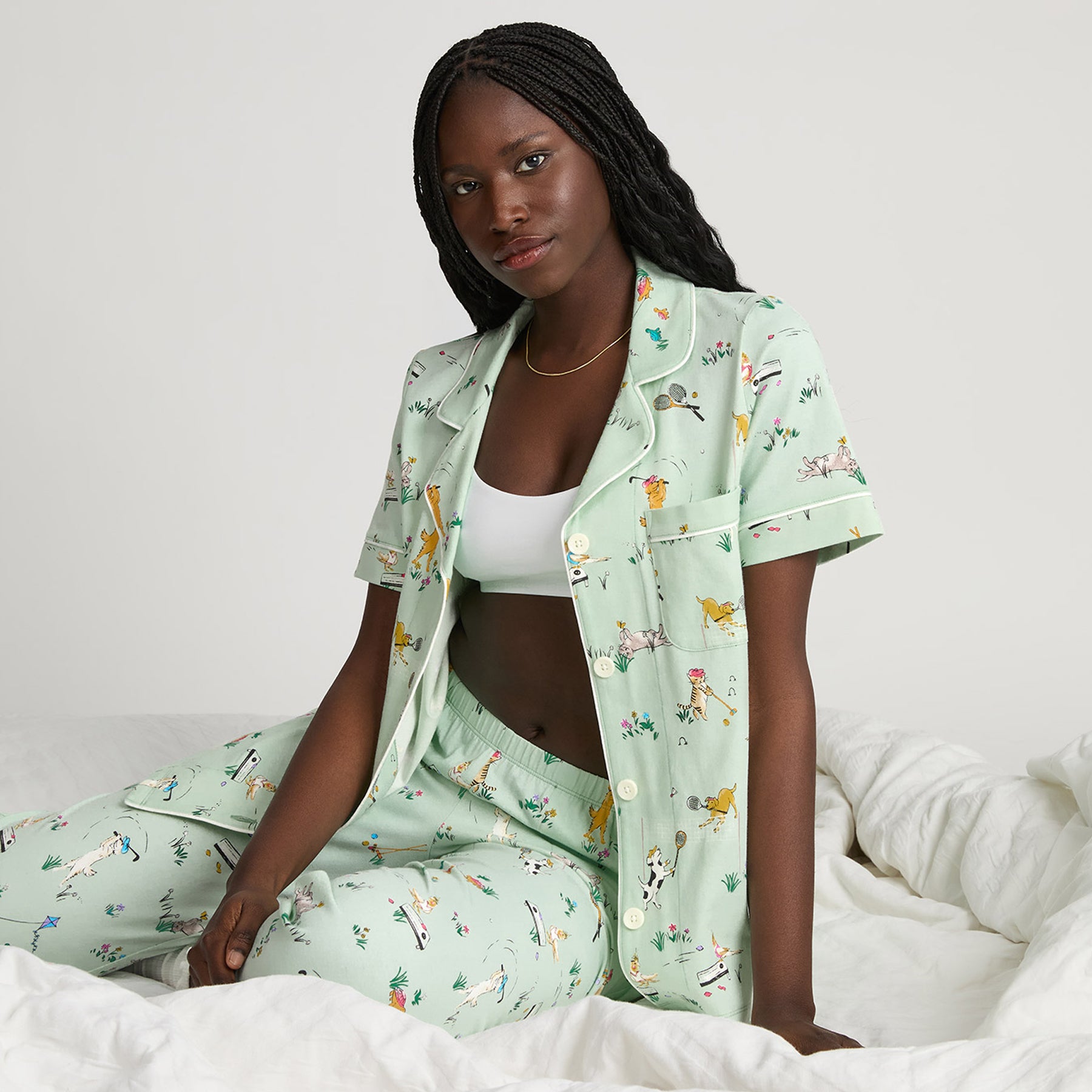 Model sitting on a bed wearing a mint green printed short sleeve pajama set with button-down top and cropped pants featuring playful dog and tennis print.