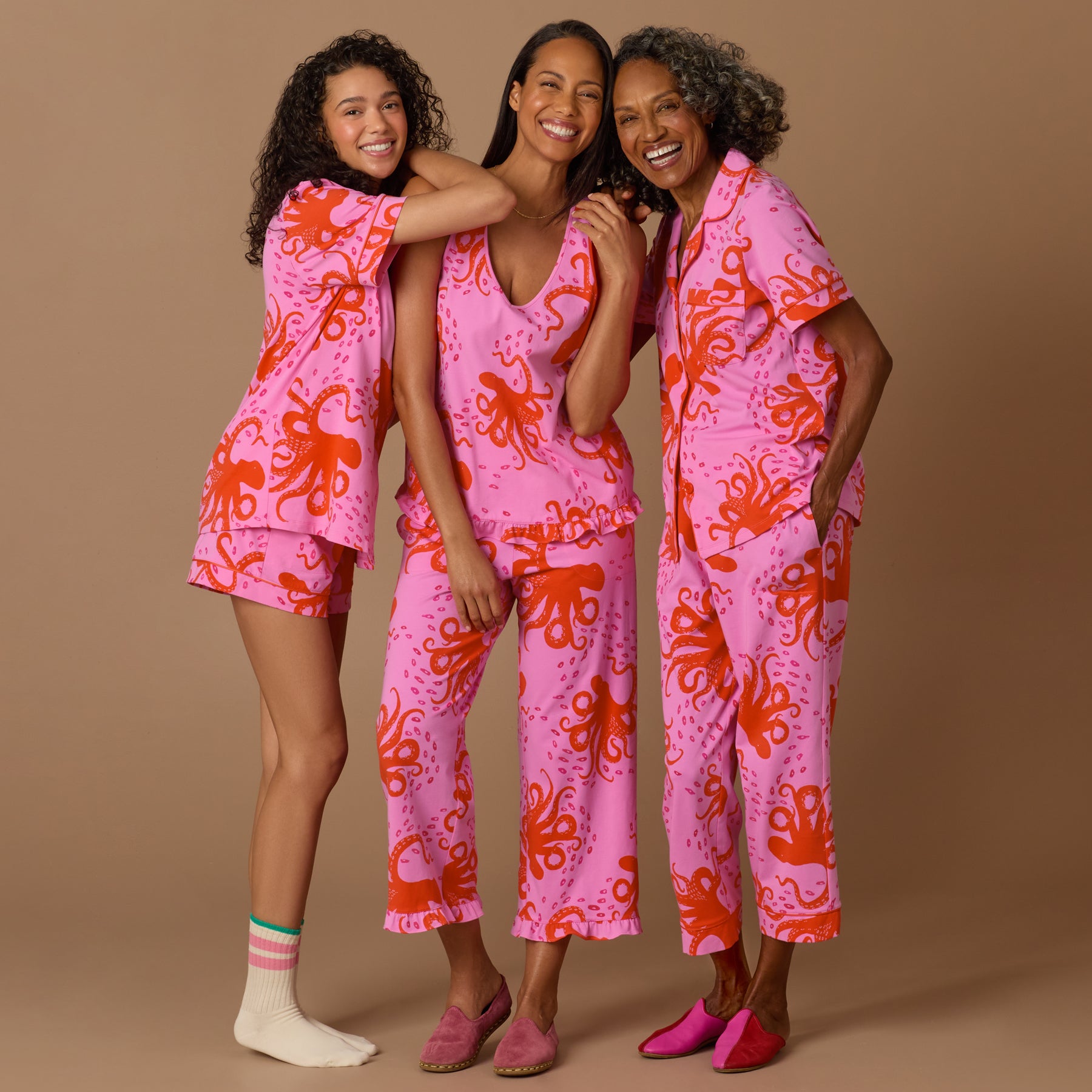 Three women of different ages wearing matching pink and coral octopus print pajamas, smiling and embracing against a beige background.