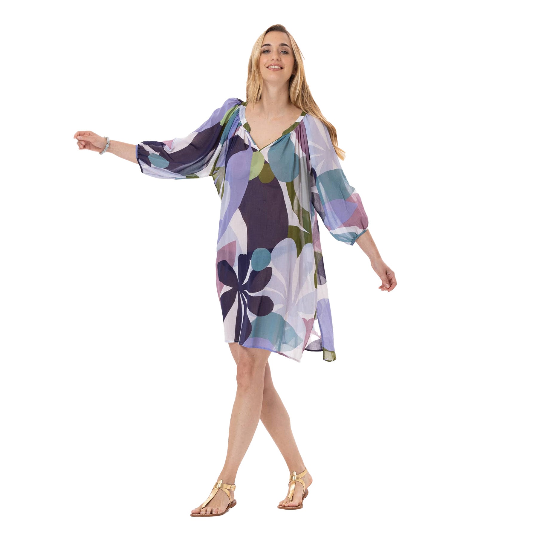Full-body view of a smiling woman wearing a loose, knee-length, multicolour floral-print cover-up dress with flowy three-quarter sleeves, walking barefoot in gold sandals.