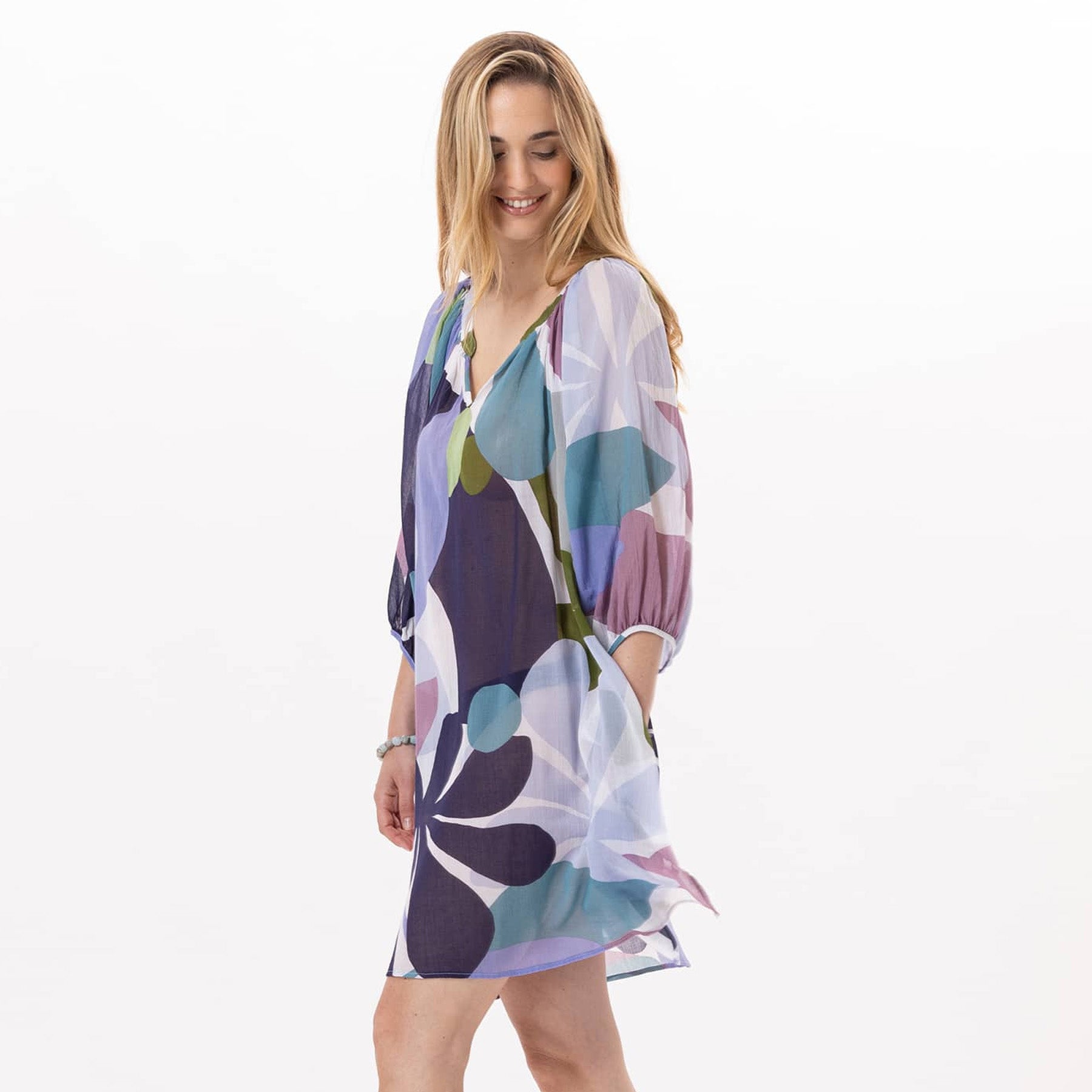 Side view of a woman smiling and looking down while wearing a sheer, flowy dress with bold floral patterns in purple, green, blue, and white.