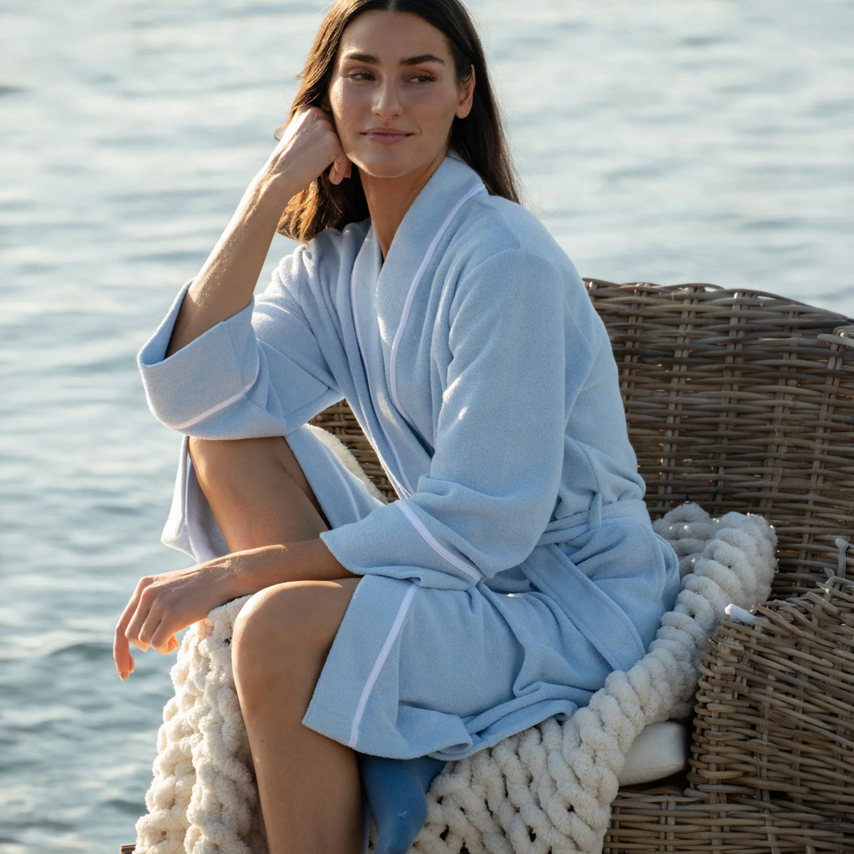 Carole Hochman light blue fleece robe with white piping, shawl collar and matching waist tie, worn by a woman relaxing in a wicker chair by the water.