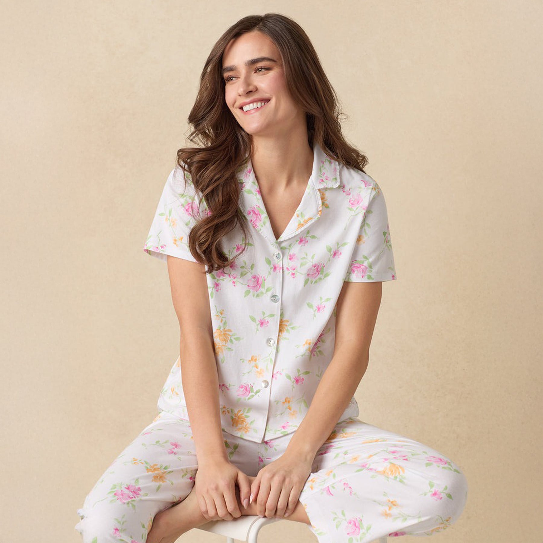 Woman sitting, smiling, in a white floral short-sleeve pajama set with a button-up top.
