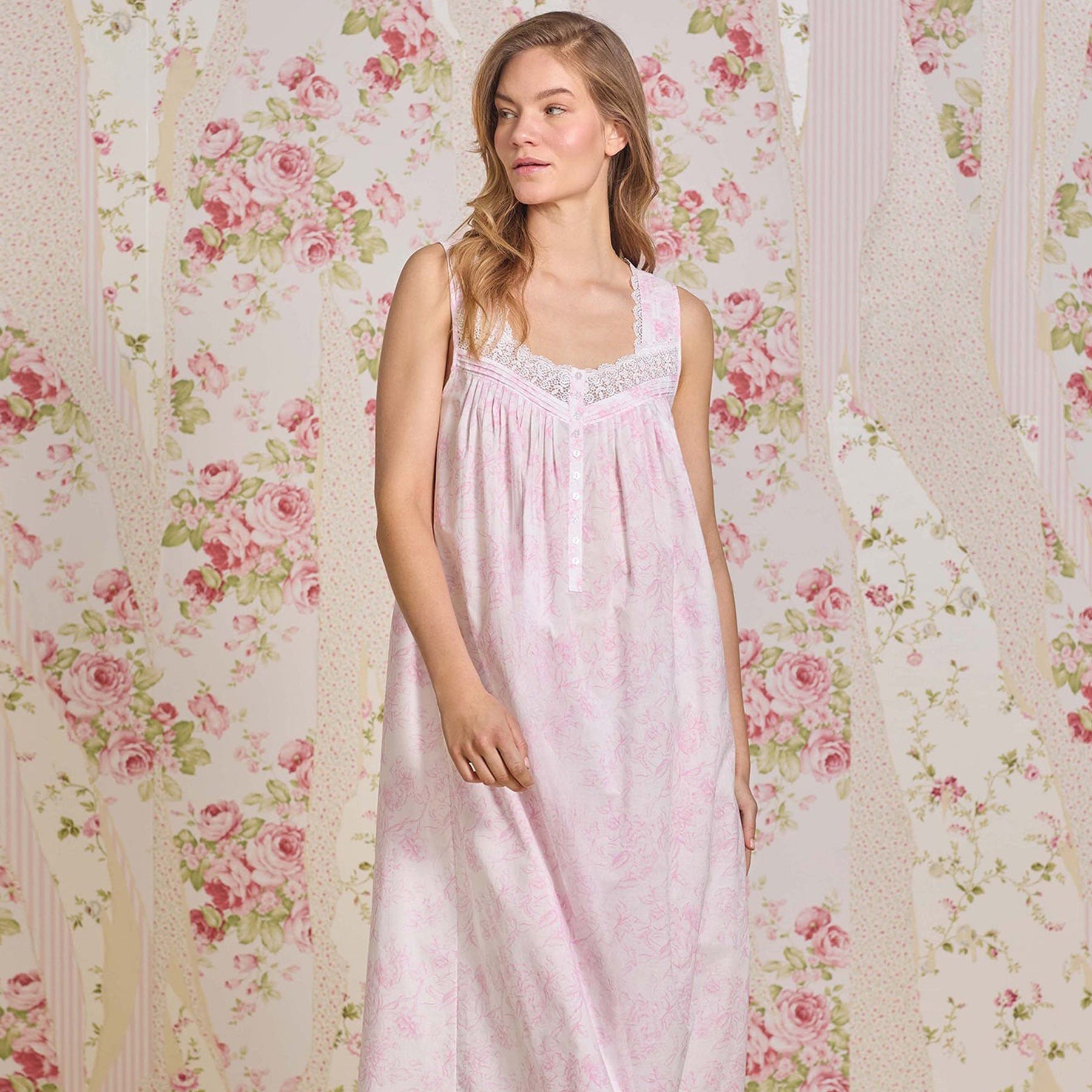 Woman standing in a pink floral cotton nightgown with lace trim, in front of a romantic floral wallpaper backdrop.