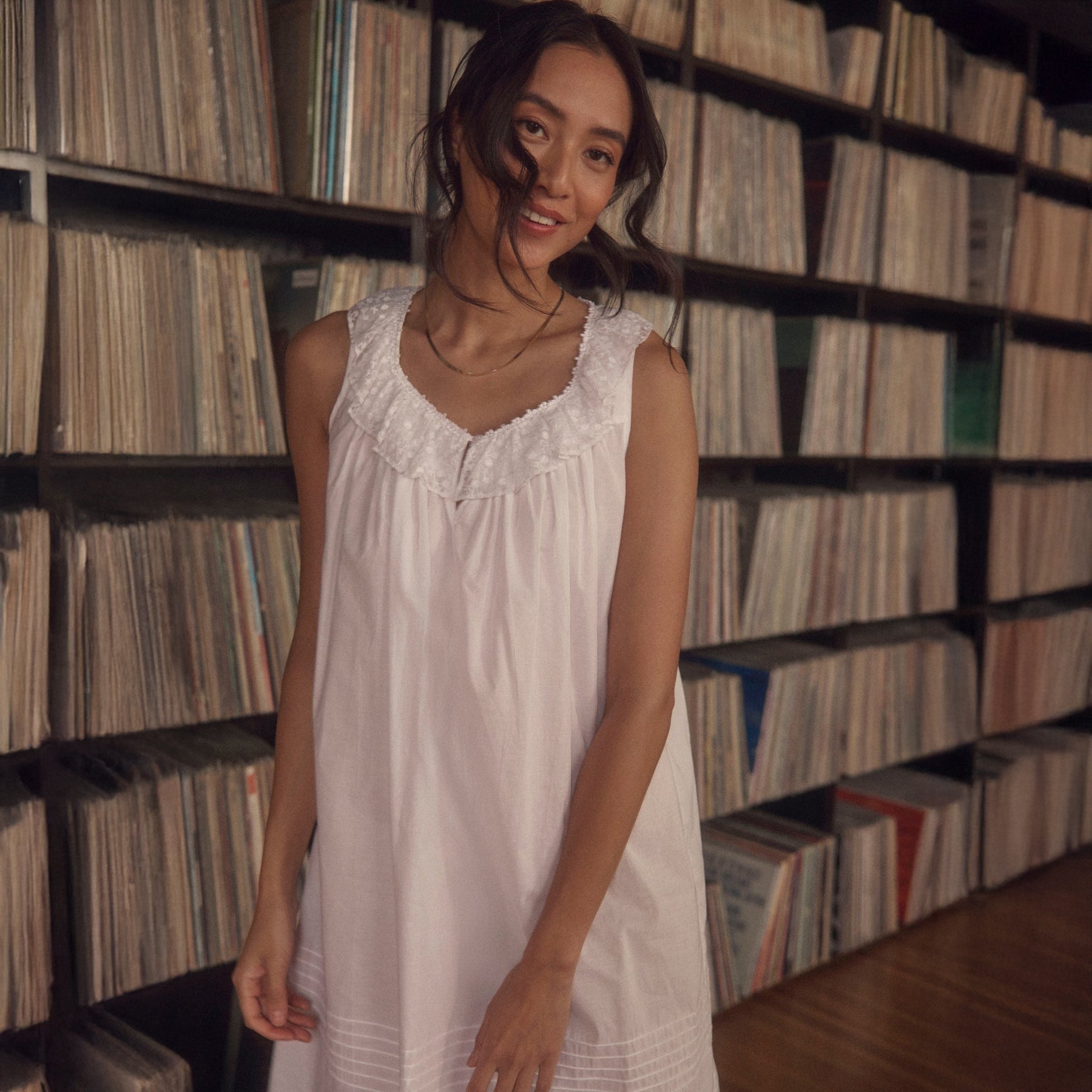 Woman in a white sleeveless nightgown with ruffled neckline, standing in front of a wall of vinyl records.
