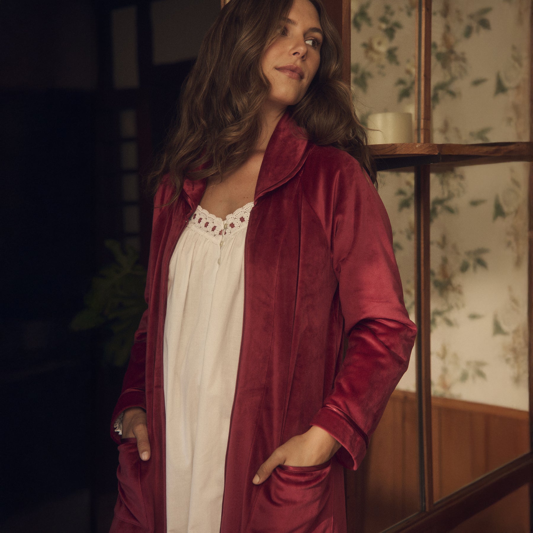 Woman wearing wine red velour robe over white nightgown, standing by a window.