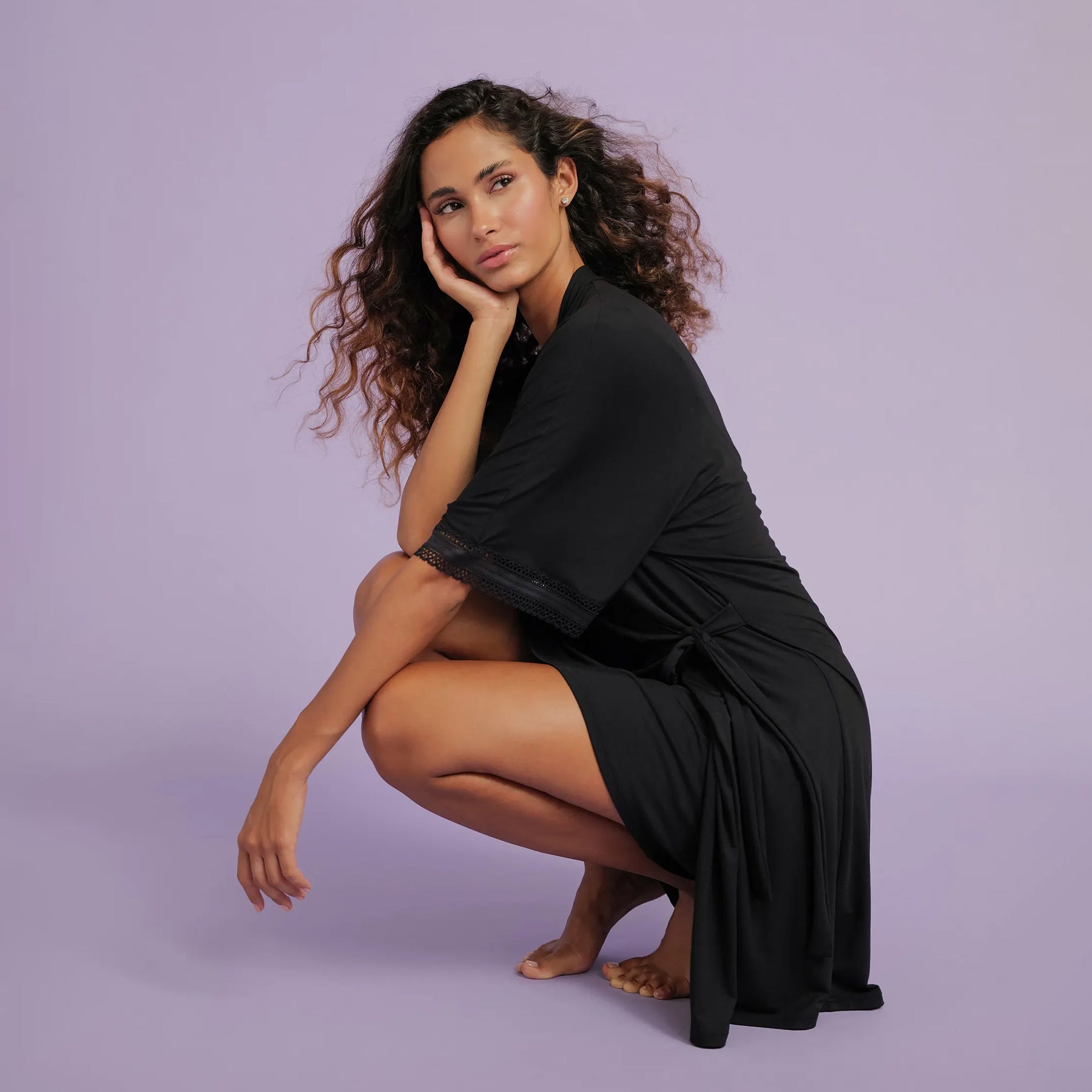 A model wearing the black robe, sitting against a soft purple background, showcasing its elegant drape and feminine details.