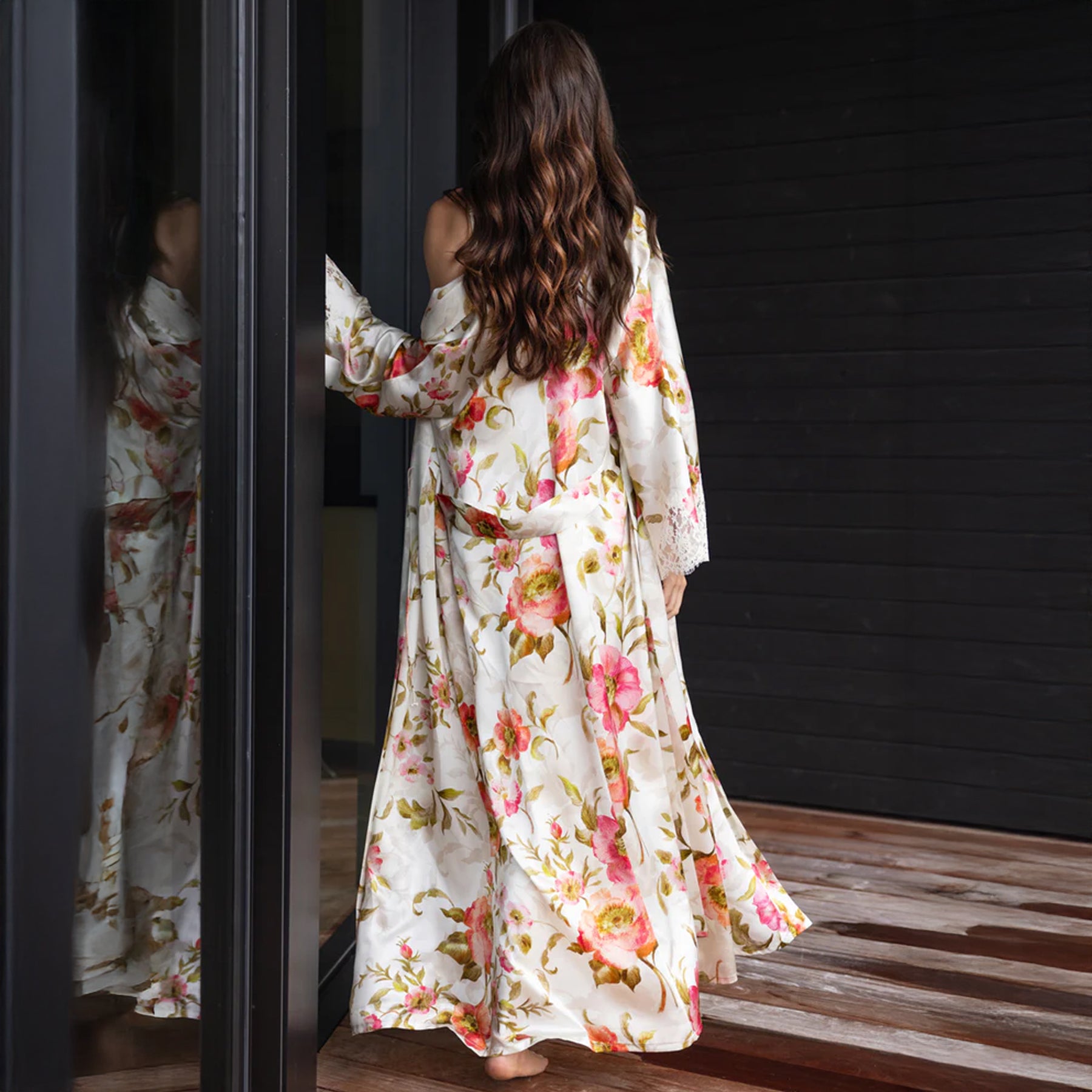 Woman wearing the silk floral robe off one shoulder, standing barefoot near a window.