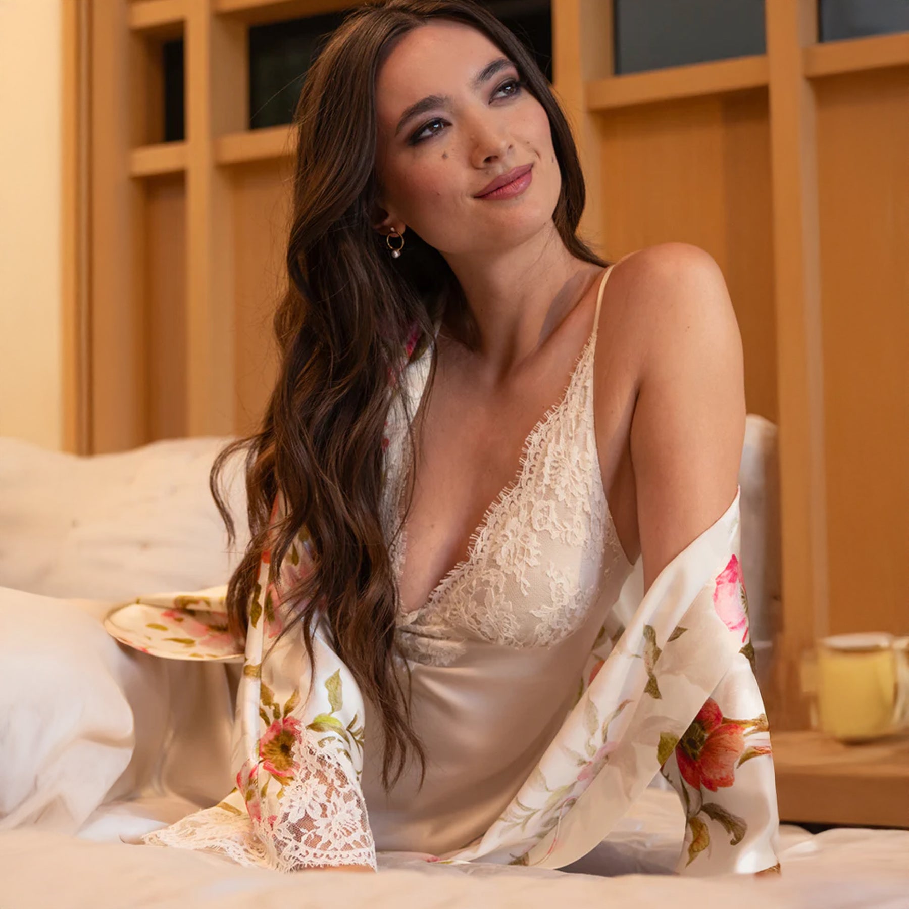 Woman seated in bed wearing a lace-trimmed silk gown under the floral robe draped off her shoulders.