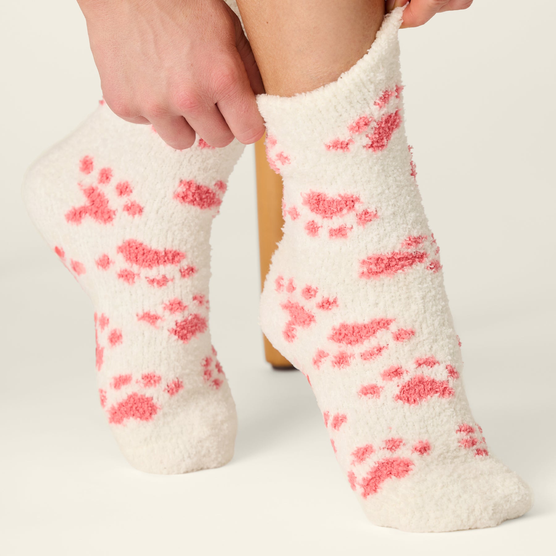 Ivory plush crew socks with pink paw prints, shown being pulled on for a cozy fit.