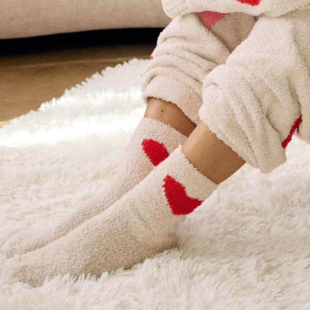 Model wearing Splendid oatmeal-coloured fuzzy crew socks with red heart accents, relaxing on a soft white faux fur rug in matching cozy loungewear.