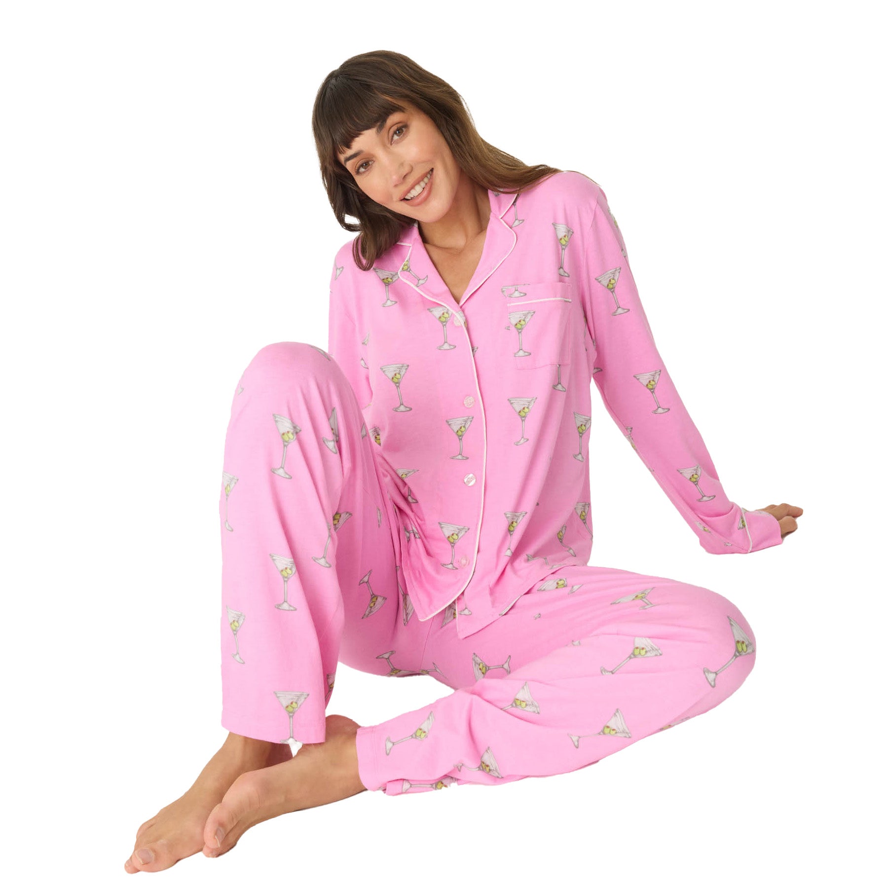 A woman sitting on the floor, smiling while wearing a pink pajama set with a martini glass print and white piping details.