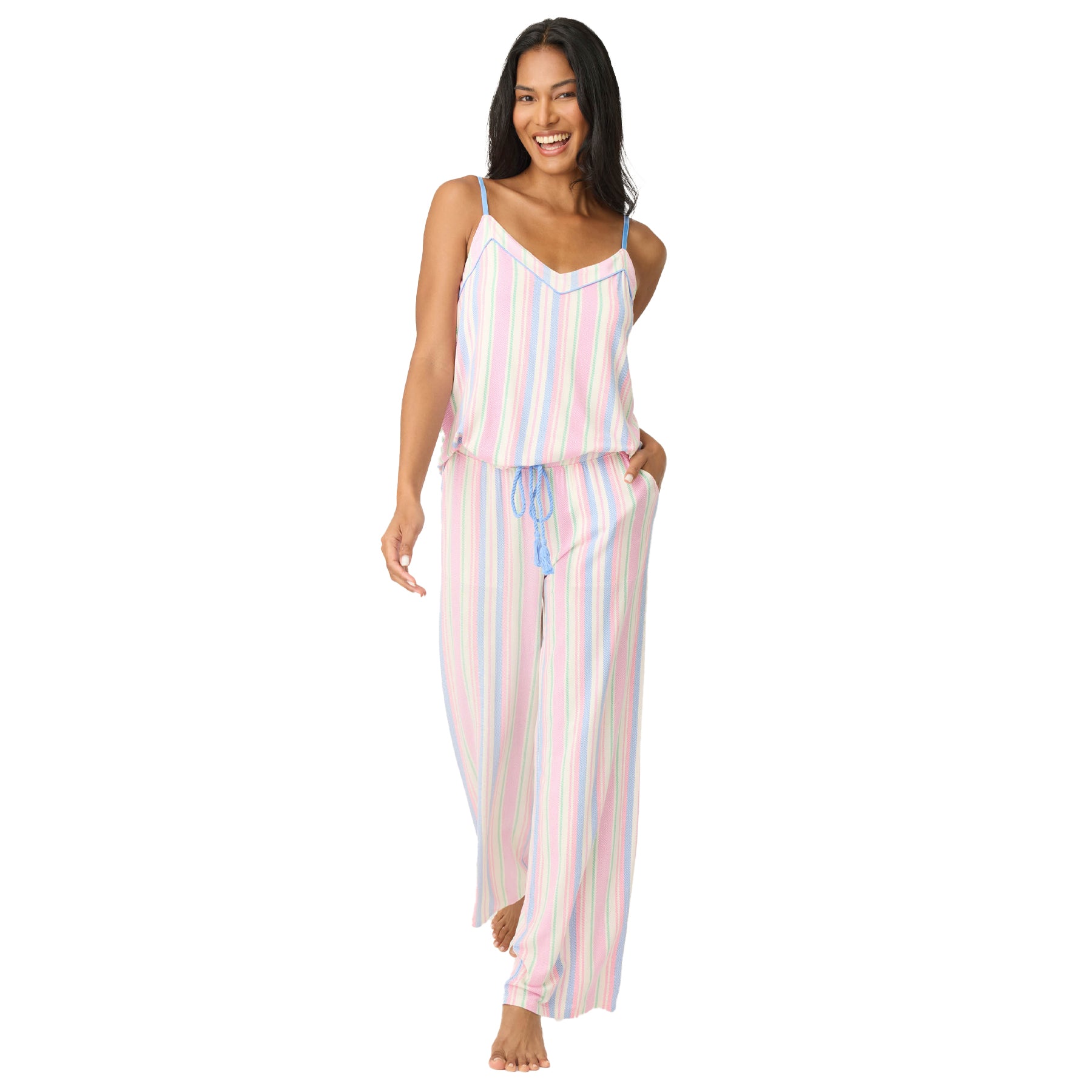Woman wearing pastel striped pyjama set with adjustable spaghetti straps and wide-leg pants, smiling and walking barefoot.