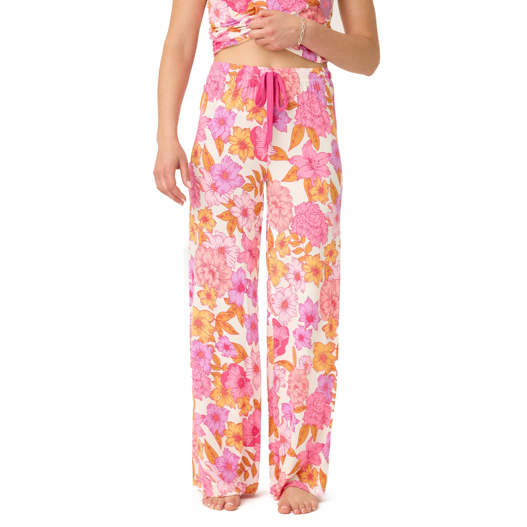 Woman wearing wide-leg pyjama pants with a vivid floral print in pink, orange, and purple tones on a natural background, featuring a bright pink drawstring waistband.