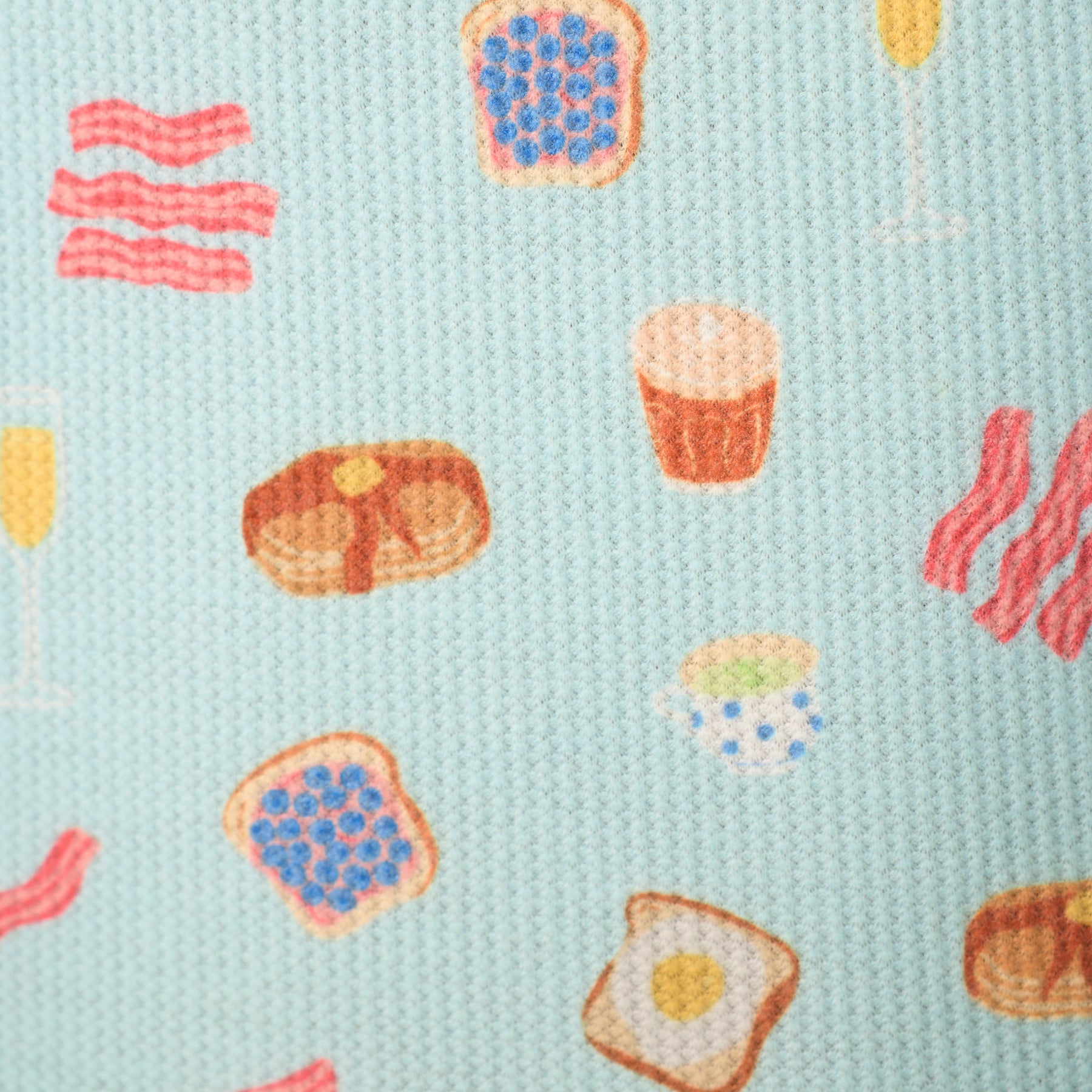Close-up of light blue waffle knit fabric with playful breakfast print featuring bacon, toast, pancakes, eggs, coffee, and champagne glasses.