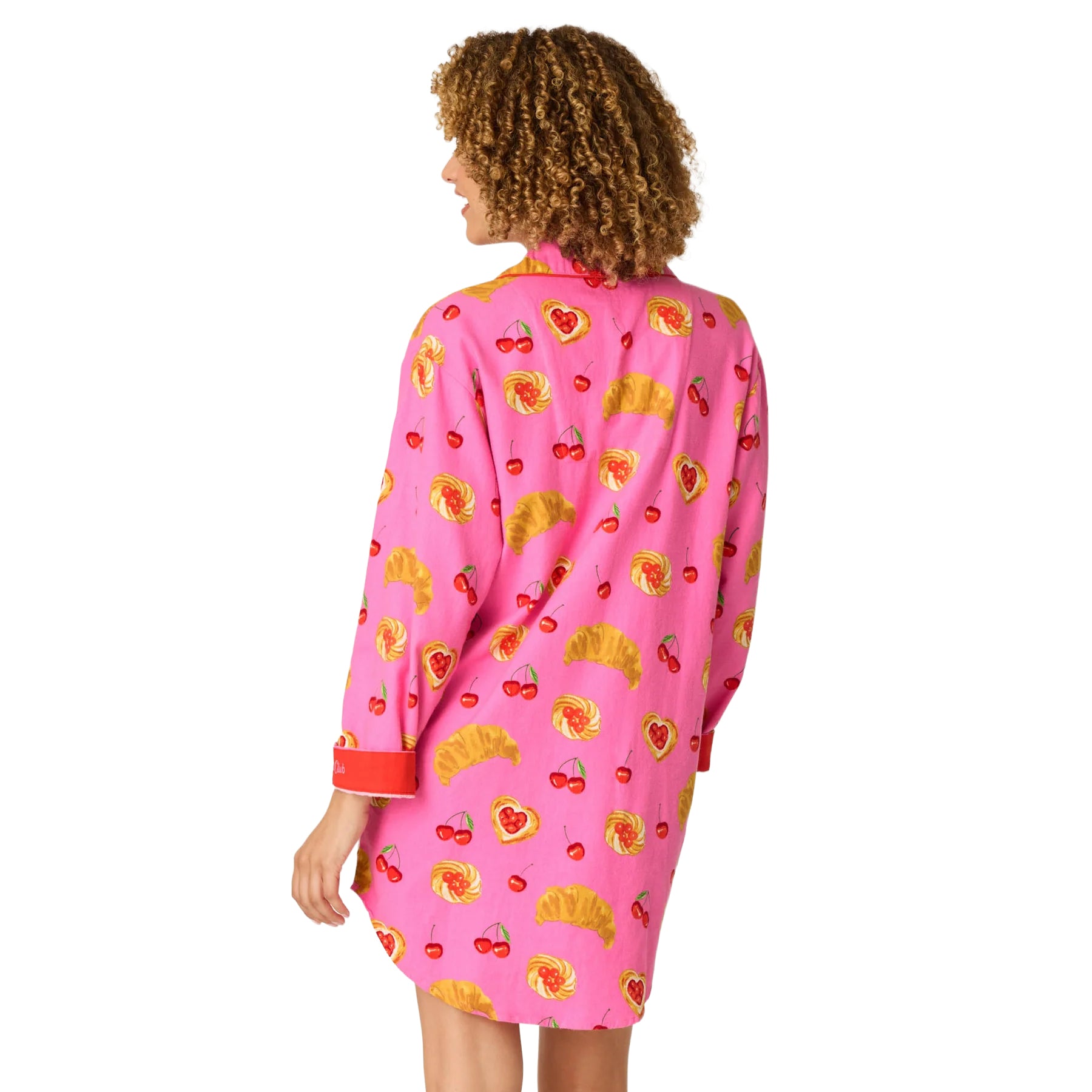 A woman shown from the back wearing a pink long-sleeve pajama shirt with a repeating print of yellow donuts, red cherries, and red hearts, against a plain white background.