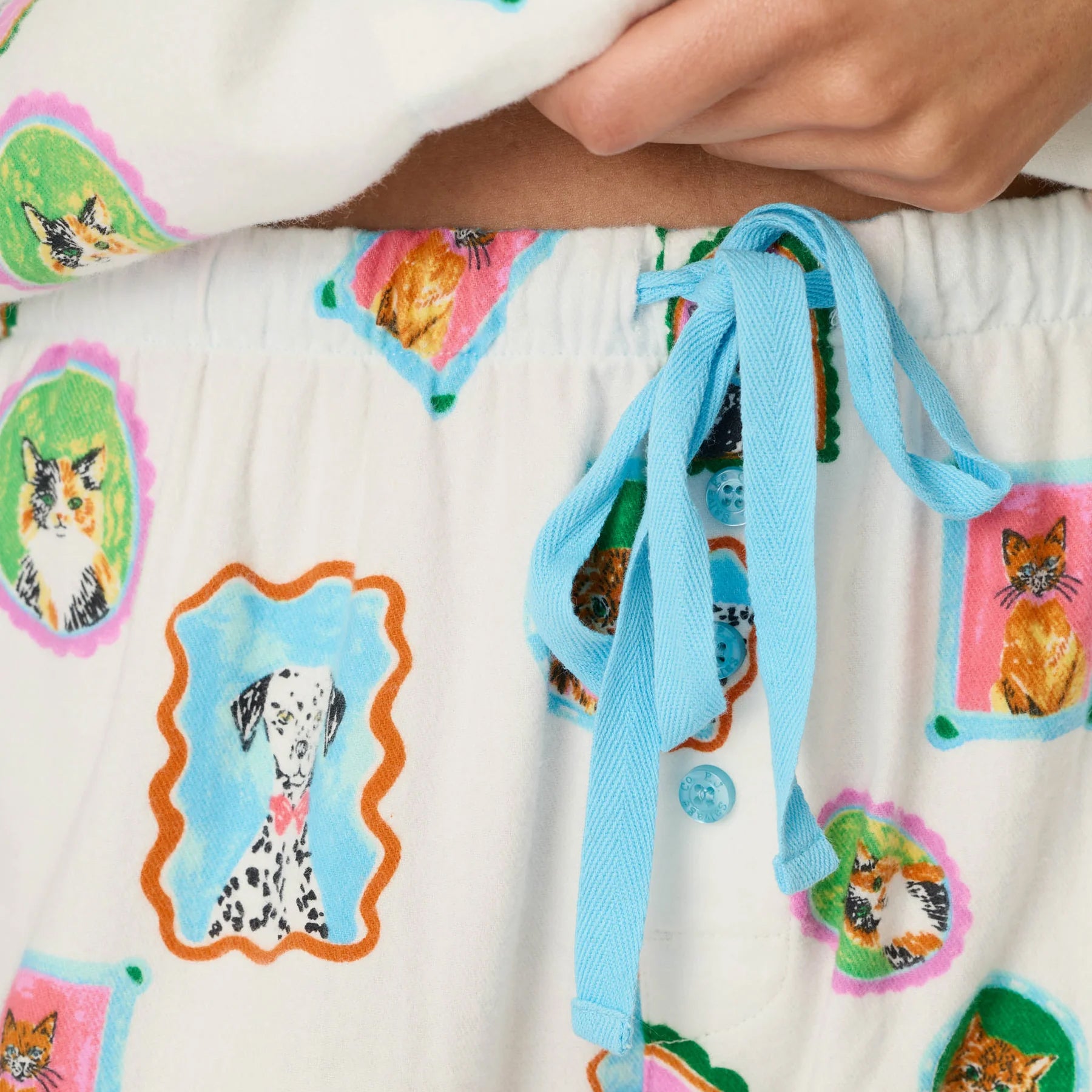 Close-up of cream pet print pajama pants featuring an elastic waistband, blue drawstring and button fly.