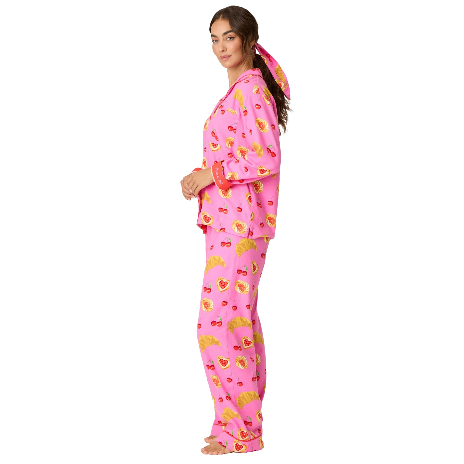 Side view of woman wearing bright pink cherry and croissant print pajamas with red cuff detailing and matching hair accessory.