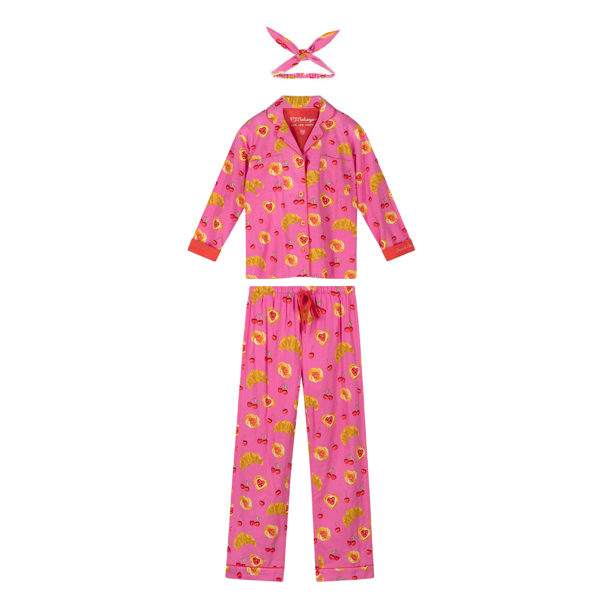 Bright pink cherry and pastry print pajama set with long-sleeve button-up top, wide-leg pants, red trim and matching hair tie.