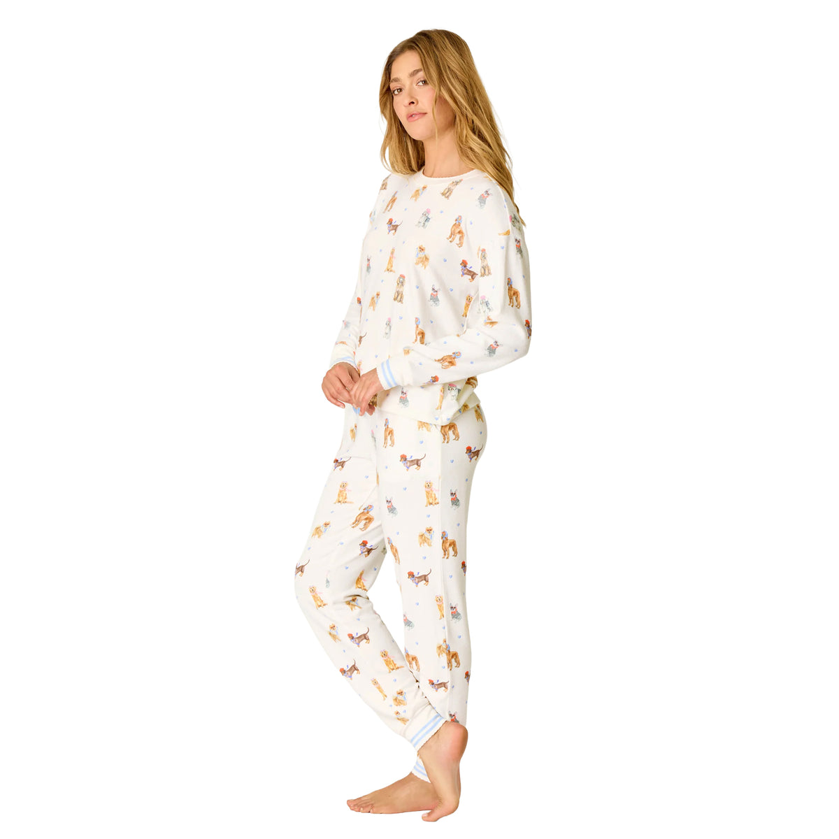 Woman wearing an ivory pajama set covered in colourful illustrated dogs, finished with soft blue striped cuffs.