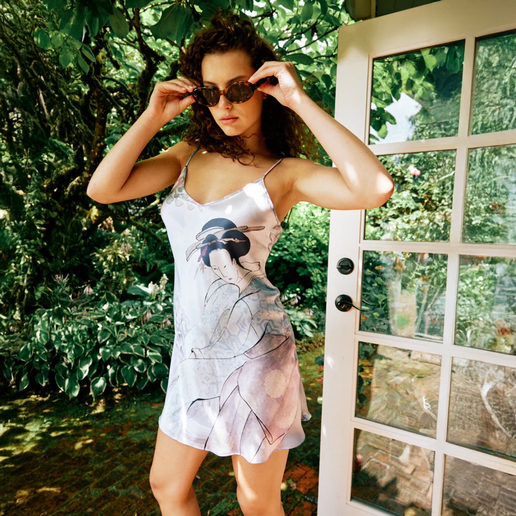 Woman wearing printed silk slip dress outdoors in garden setting, styled with sunglasses for a chic summer look