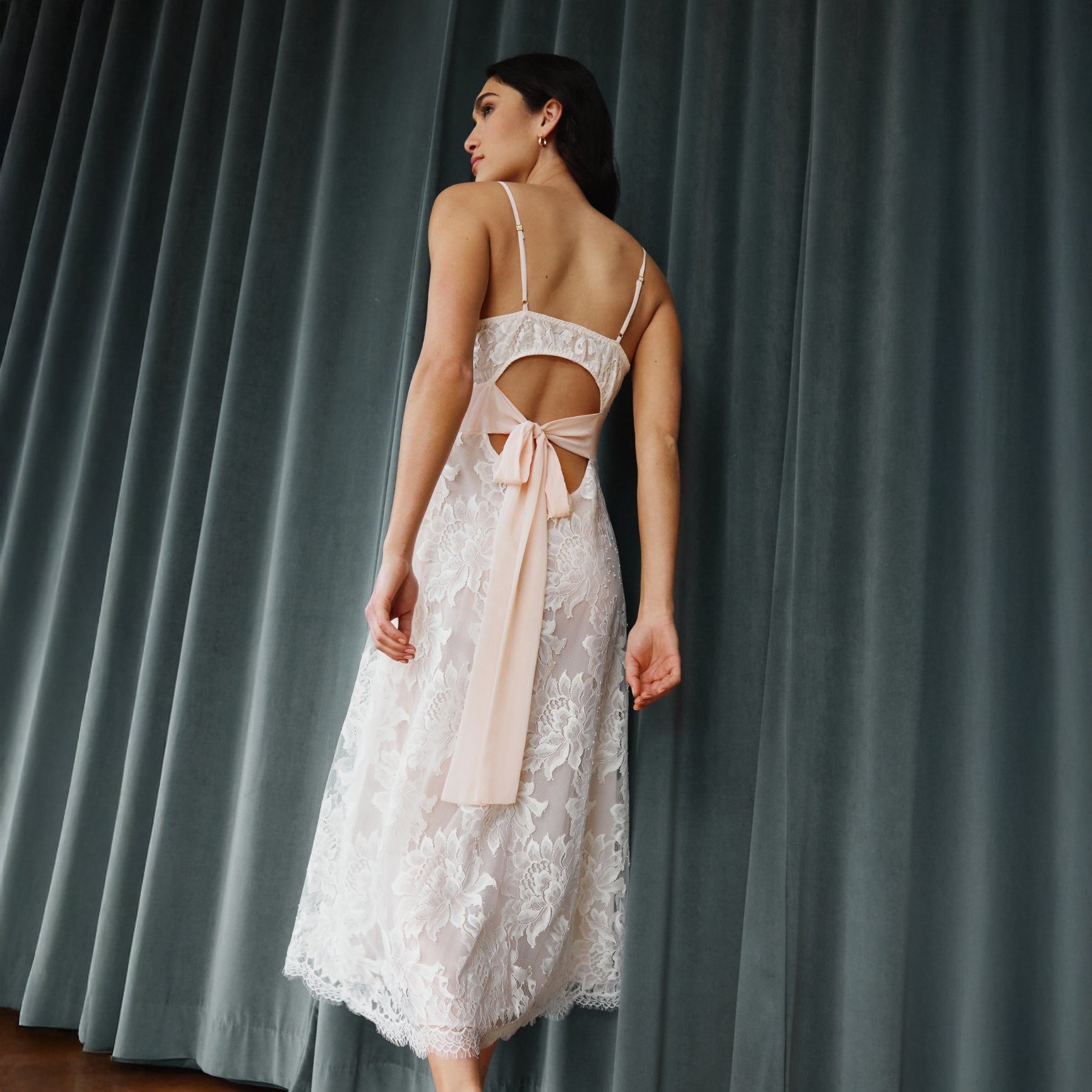 Model wearing a blush lace bridal gown with open back and soft chiffon tie detail, photographed from the back against a draped curtain backdrop.