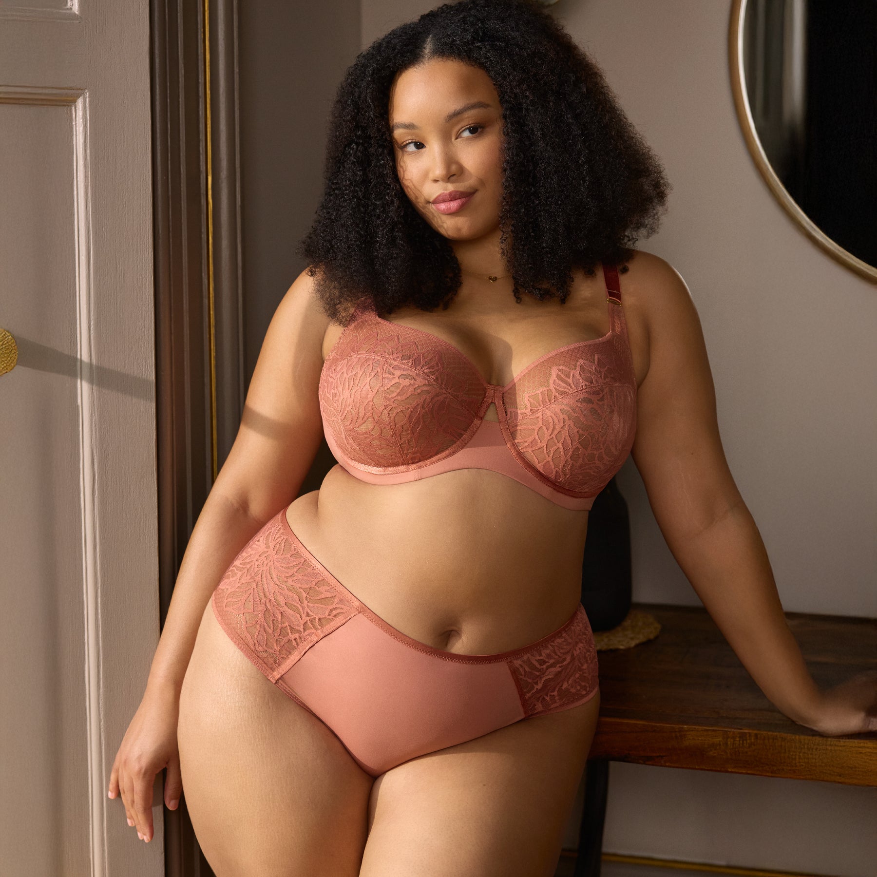 Woman wearing sienna rose lace bra and matching full brief, leaning against a wooden table.
