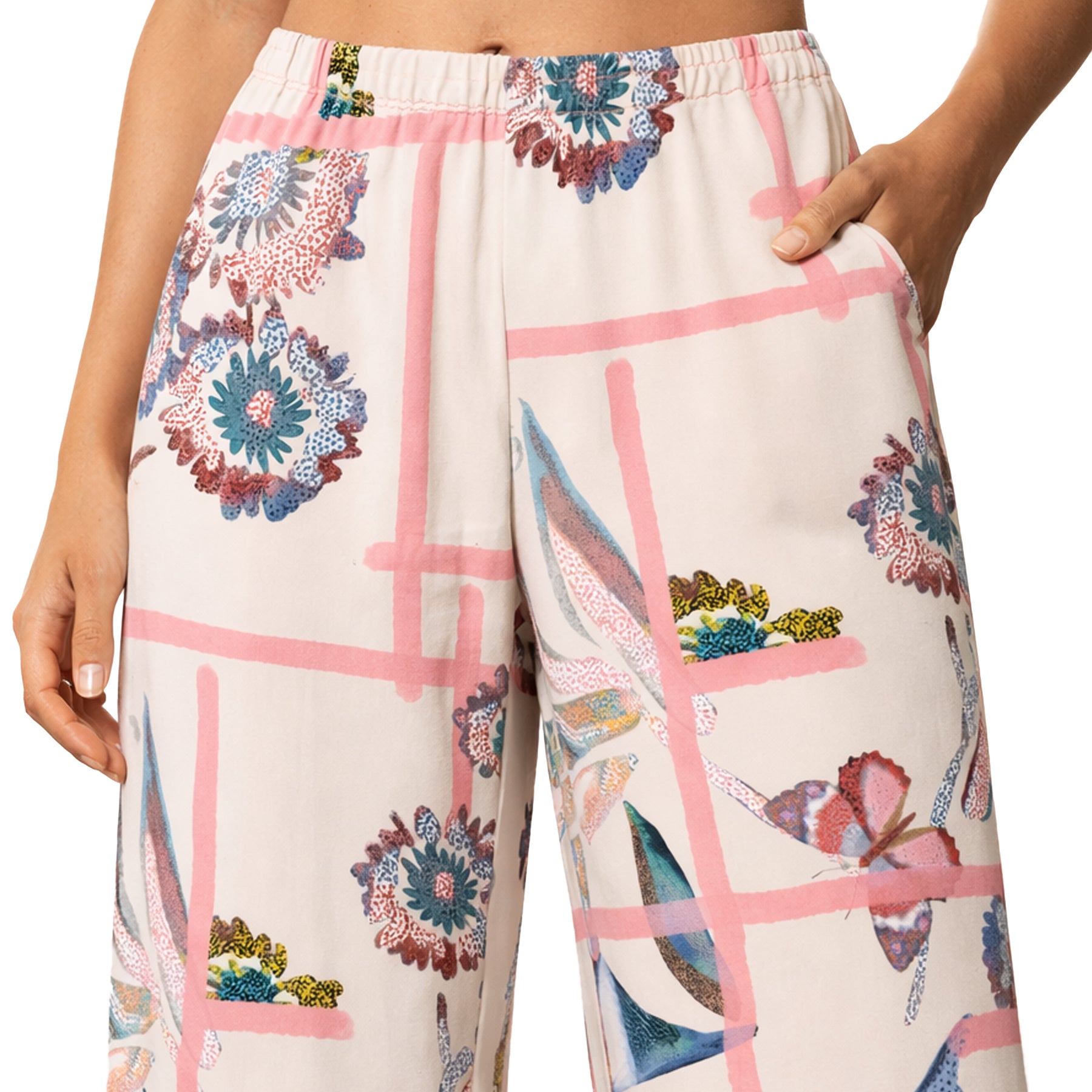 Close-up view of a woman wearing full-length pyjama pants in a cream tone with fragmented garden print, featuring pink grid accents, floral and bird motifs, elastic waistband without ties, and decorative geometric cuffs at the hem.