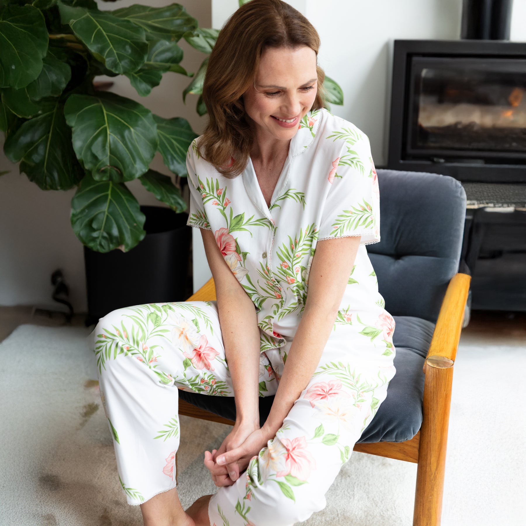 Women’s floral cotton pyjama set in a lifestyle setting, seated indoors, lightweight white fabric with tropical pink and green print, relaxed and breathable fit