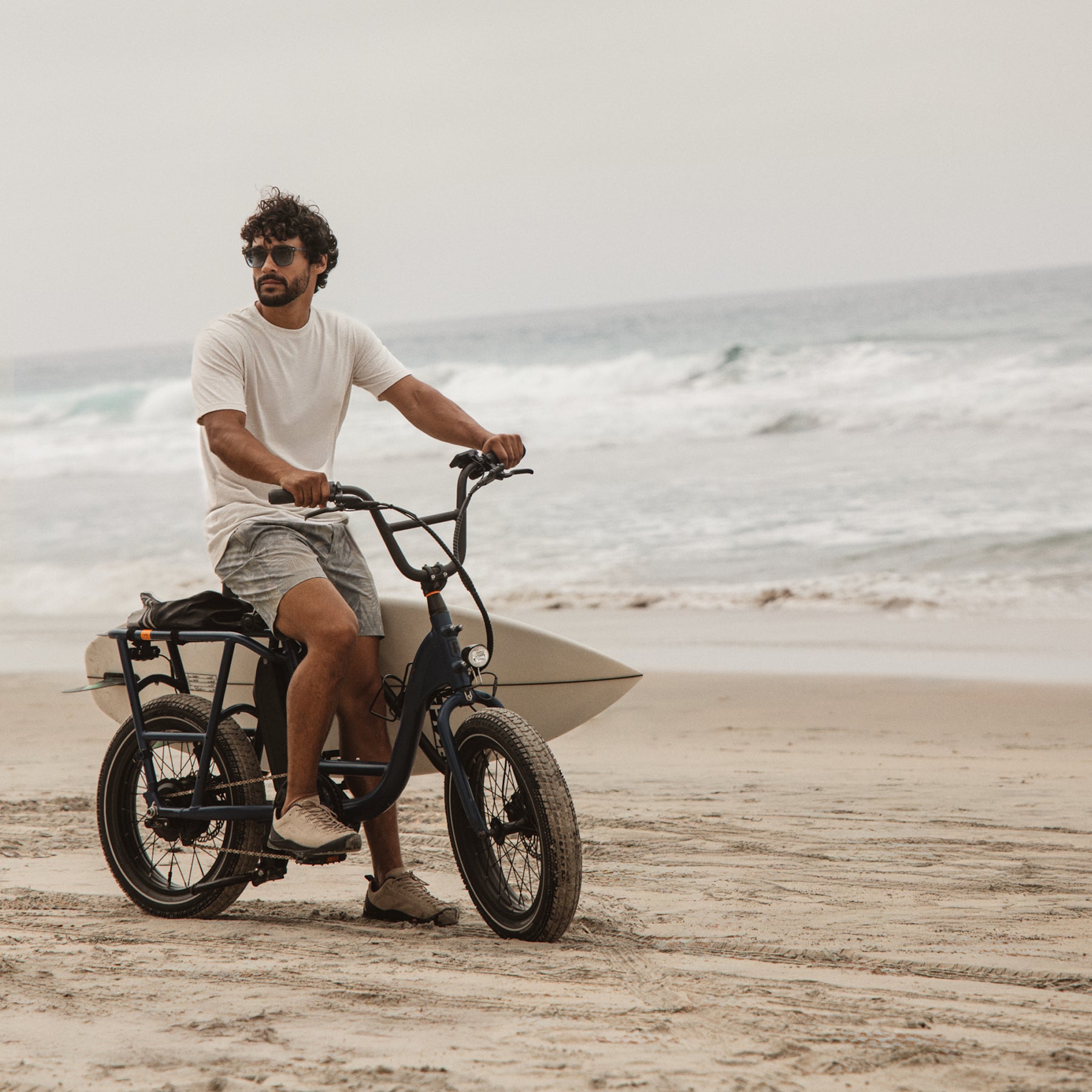 Man riding an electric bike along the beach wearing palm print swim shorts, carrying a surfboard with ocean waves in the background