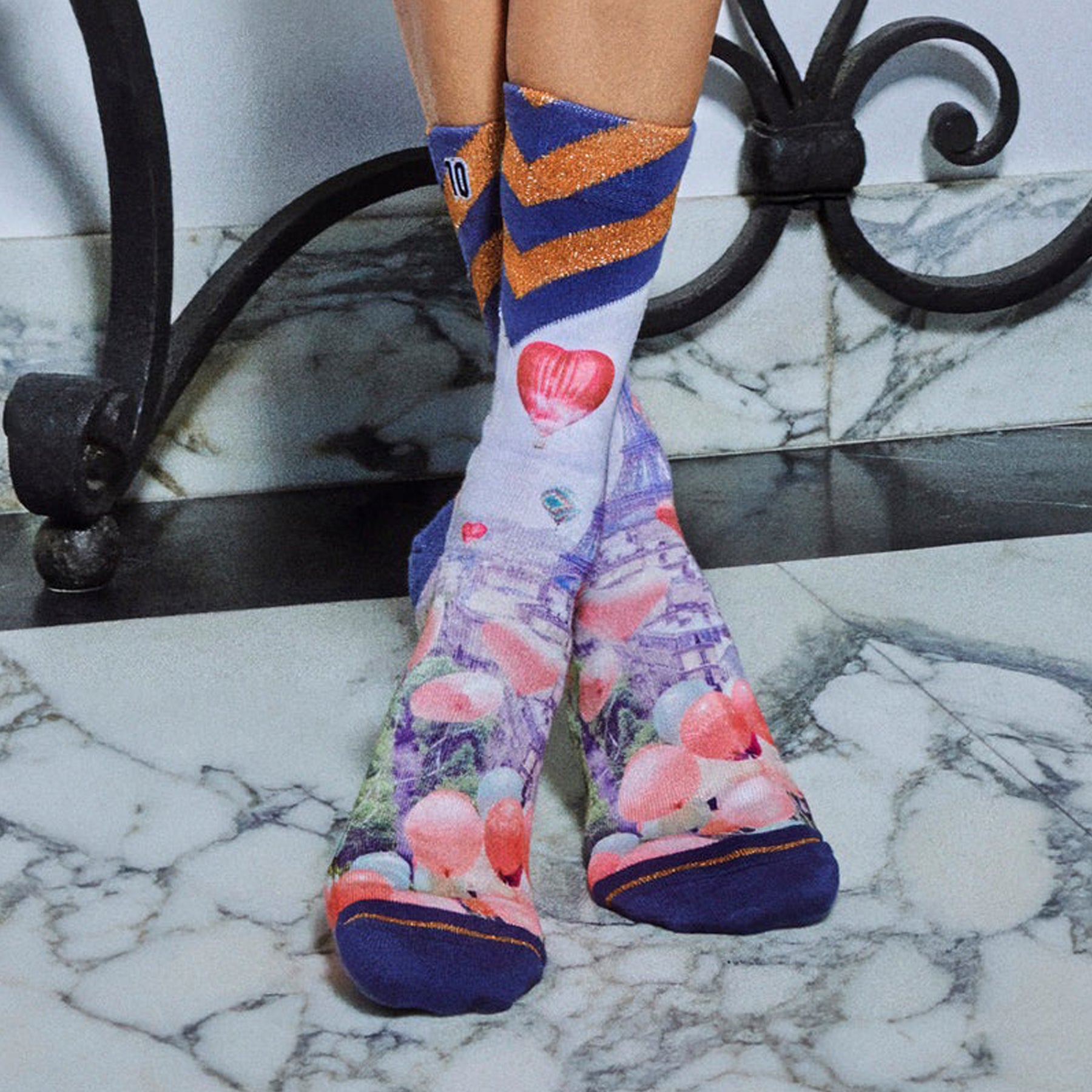 Person wearing colourful Paris-themed socks with Eiffel Tower and balloon print, standing on marble floor.