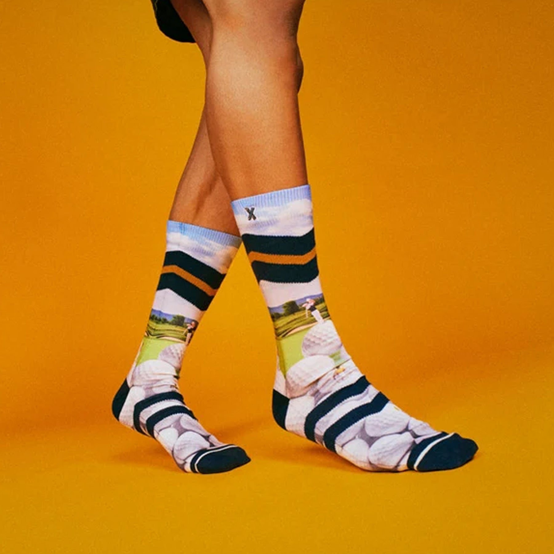 Model wearing golf-themed crew socks featuring golf balls, course scenery, and navy stripes against a bright orange background.
