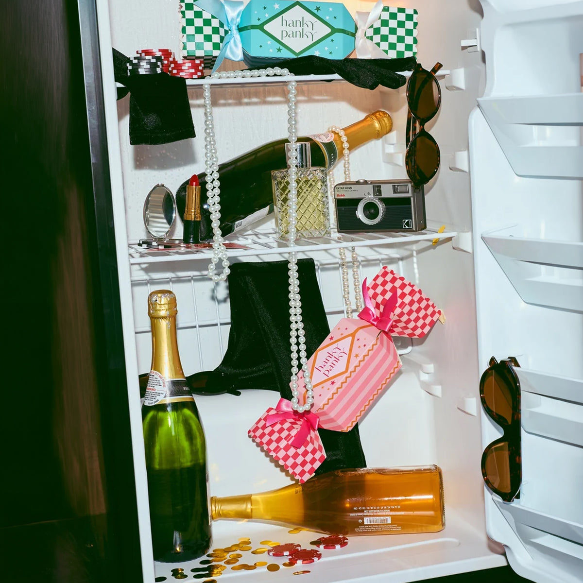 Hanky Panky holiday cracker gift boxes in blue and pink displayed inside a refrigerator alongside champagne bottles, pearls, sunglasses, and vintage accessories.