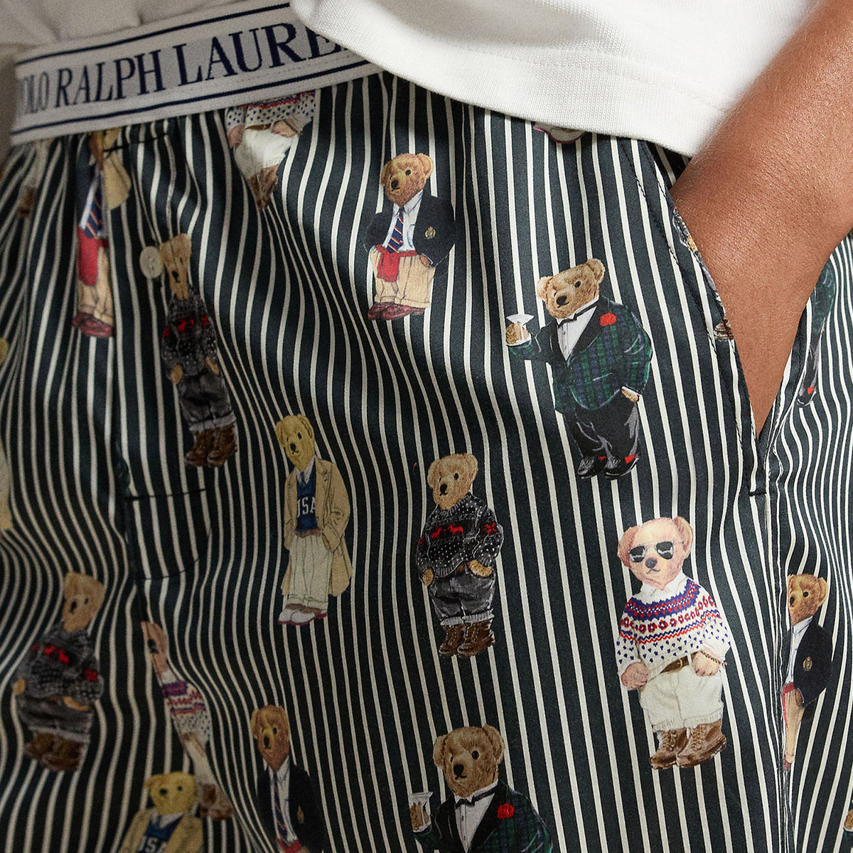 Close-up of Polo Ralph Lauren pajama pants featuring black and white vertical stripes, an all-over Polo Bear print in various outfits, and a branded elastic waistband.