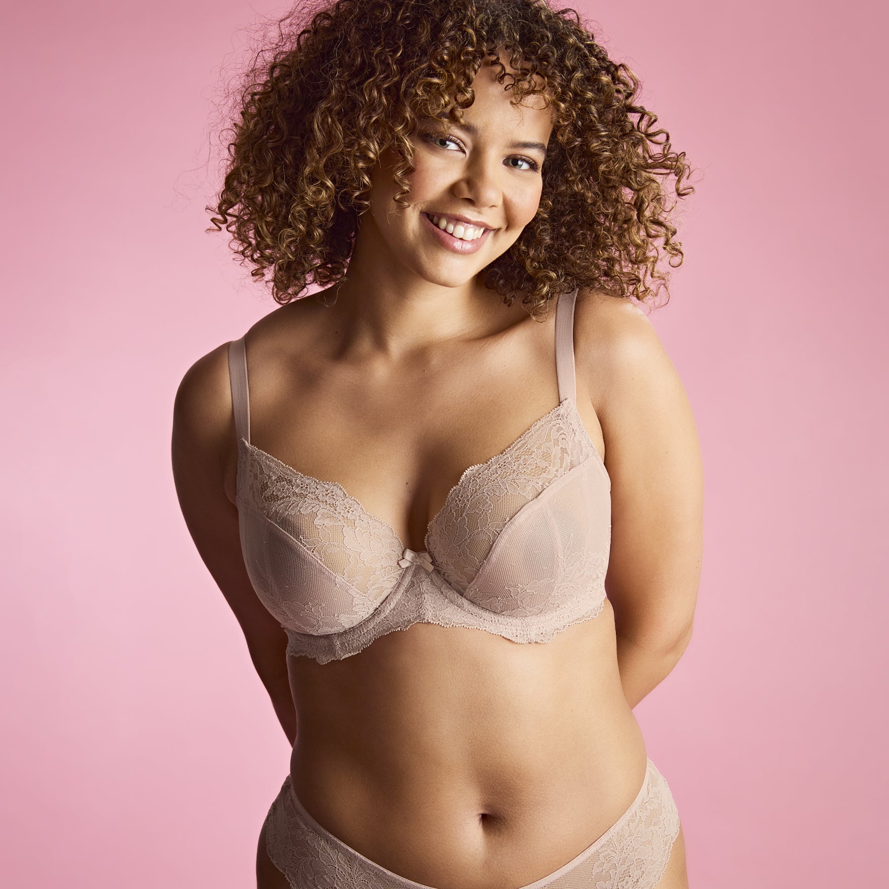 Smiling woman wearing vintage beige lace bra and brief, posing against a pink background.