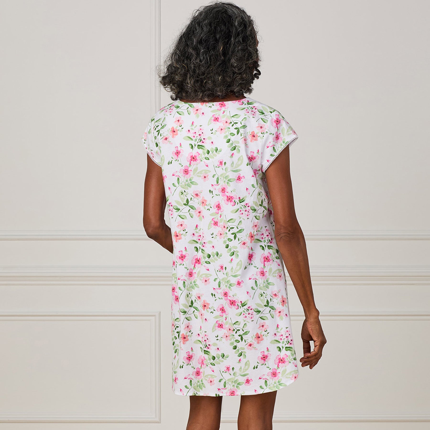 Back view of a woman wearing a white cotton nightgown with an all-over pink floral print and short sleeves.