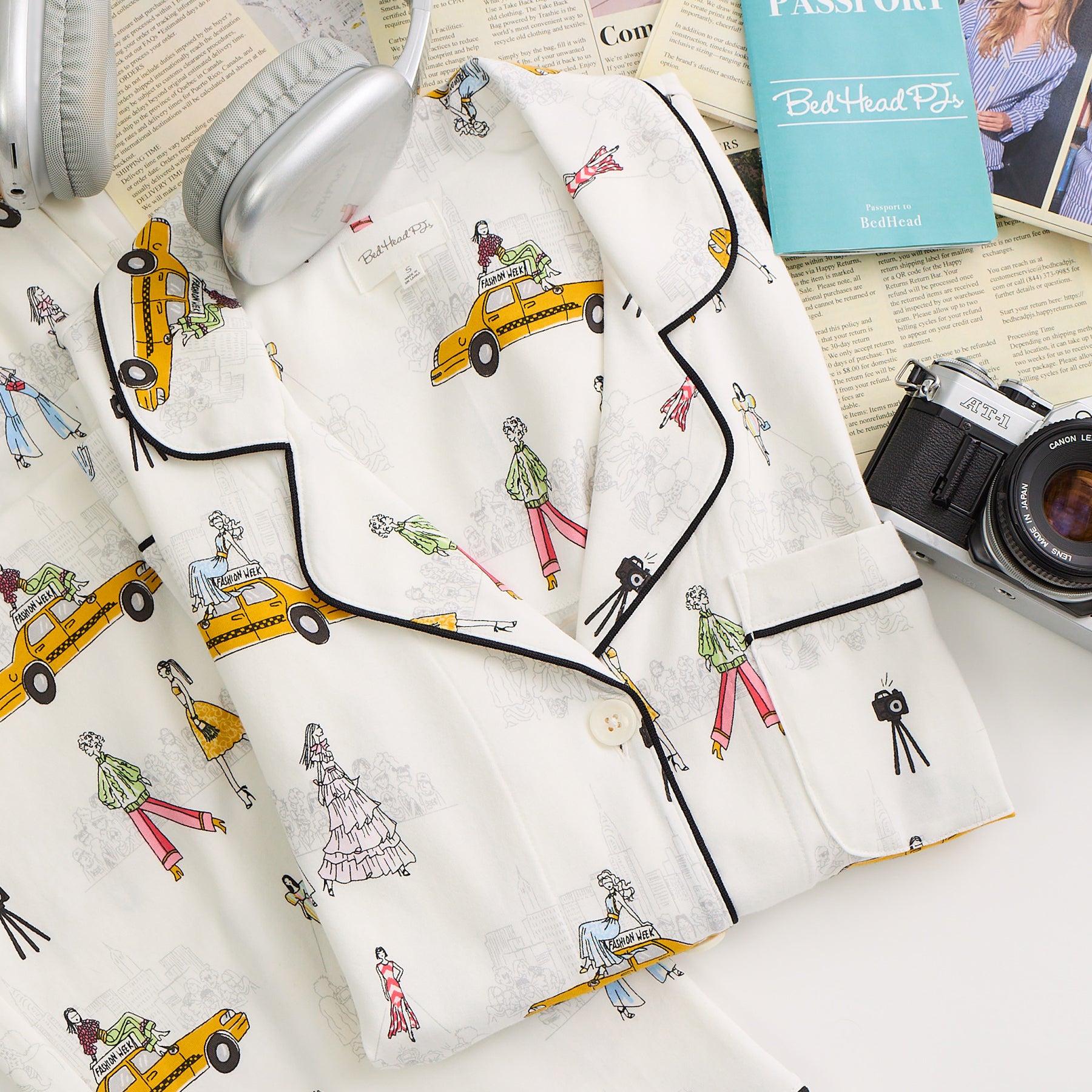 Flat lay of a women’s short sleeve pyjama top with a button front, black piping, and a fashion illustration print featuring yellow taxis and runway-inspired figures.