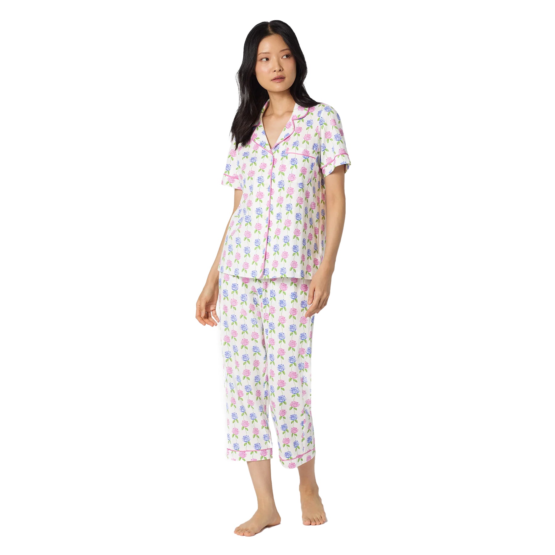 Woman wearing a white short sleeve pyjama set with pink and blue hydrangea floral print, featuring a button-front top with pink piping and cropped pants.