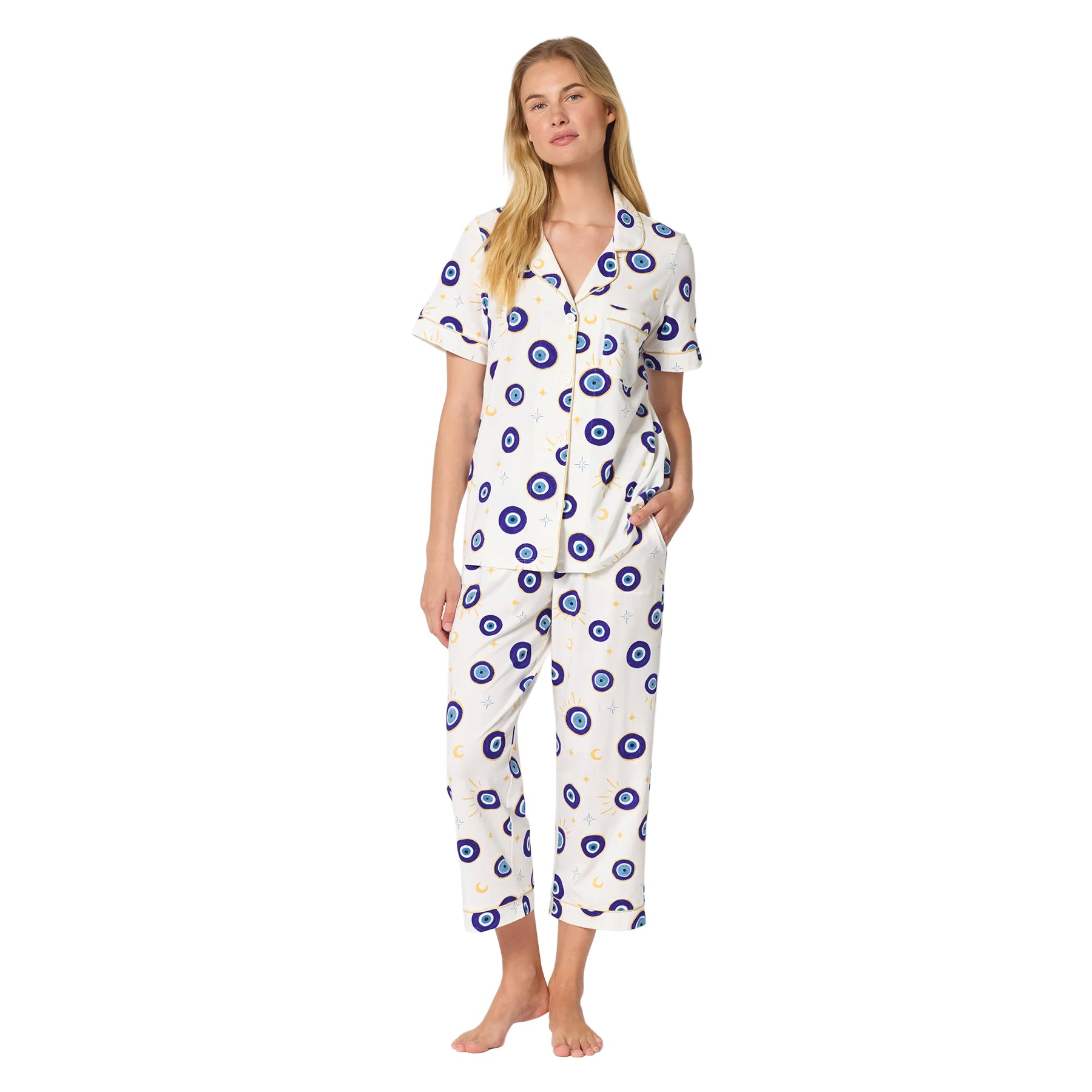 Woman wearing an ivory short-sleeve pajama set with cropped pants, featuring a blue evil eye, moon and star print with gold piping.